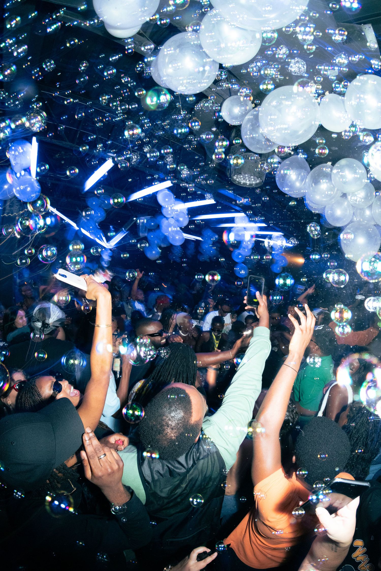 Nightclub crowd with raised hands under illuminated white balloons and blue lighting effects.
