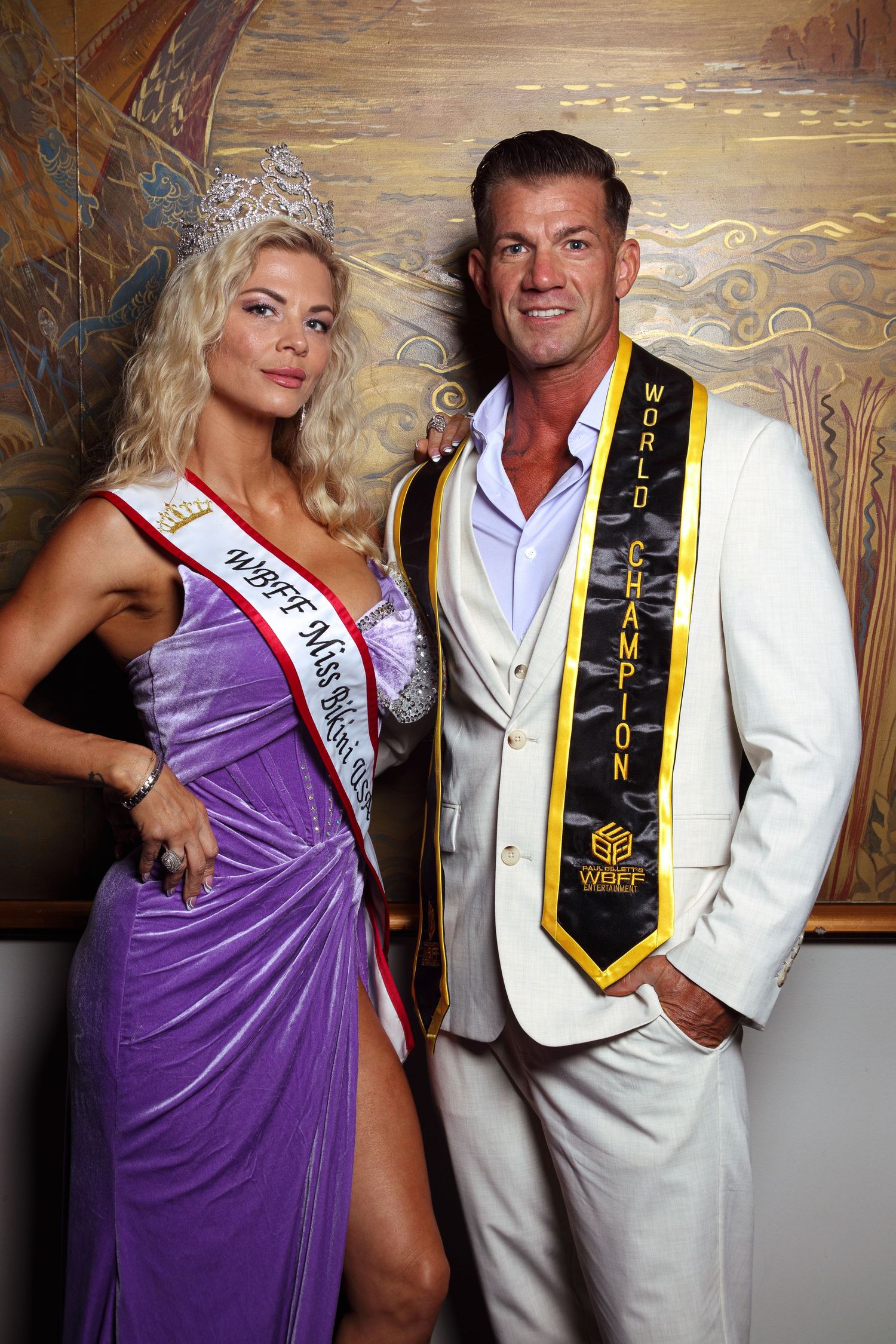WBFF World Champion man in white suit poses with Miss WBFF titleholder in purple gown and crown.