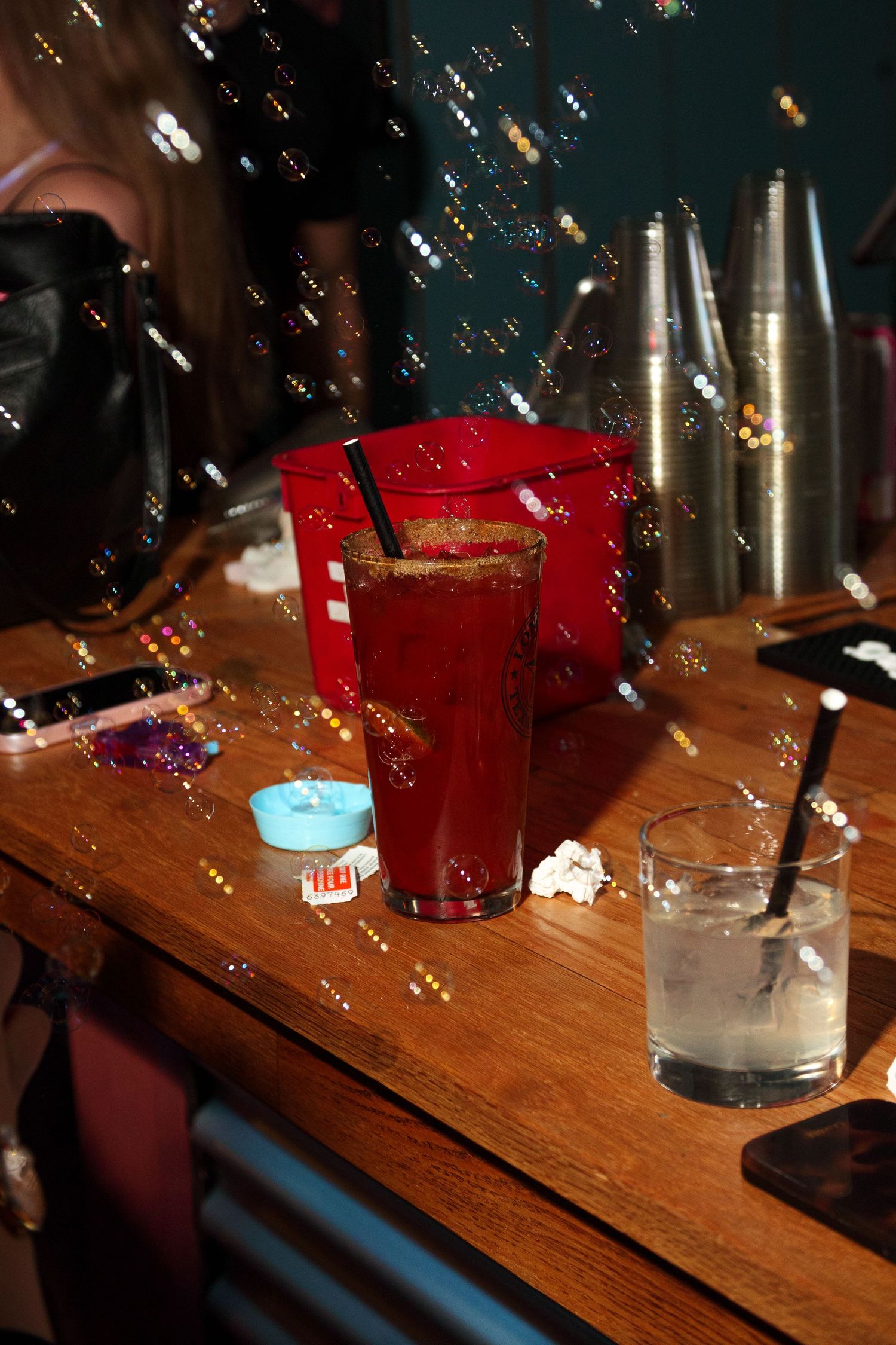 Drinks and bottles arranged on a wooden bar counter in dim lighting with glowing reflections.