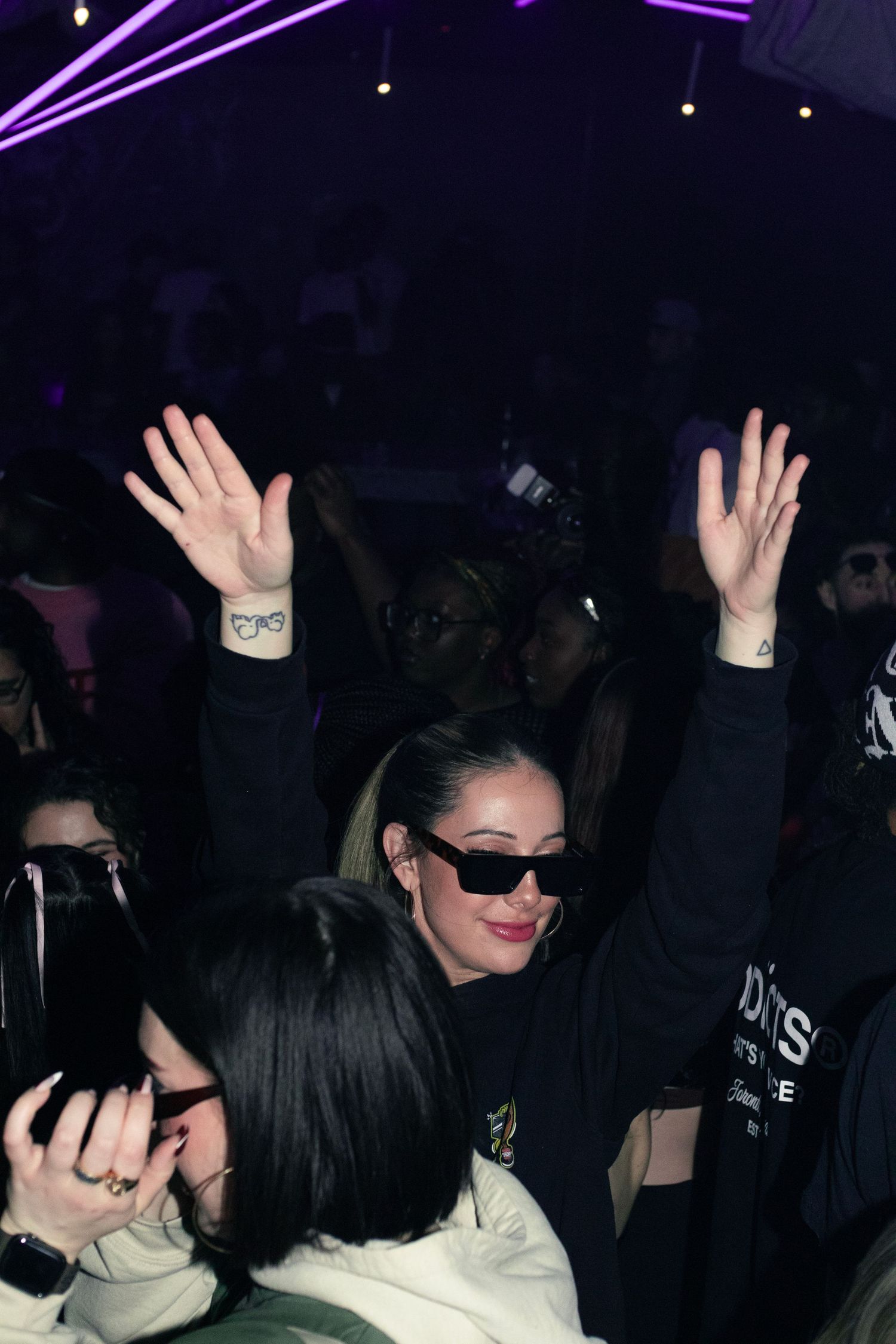 Nightclub crowd member raises hands in celebration under purple neon lighting.