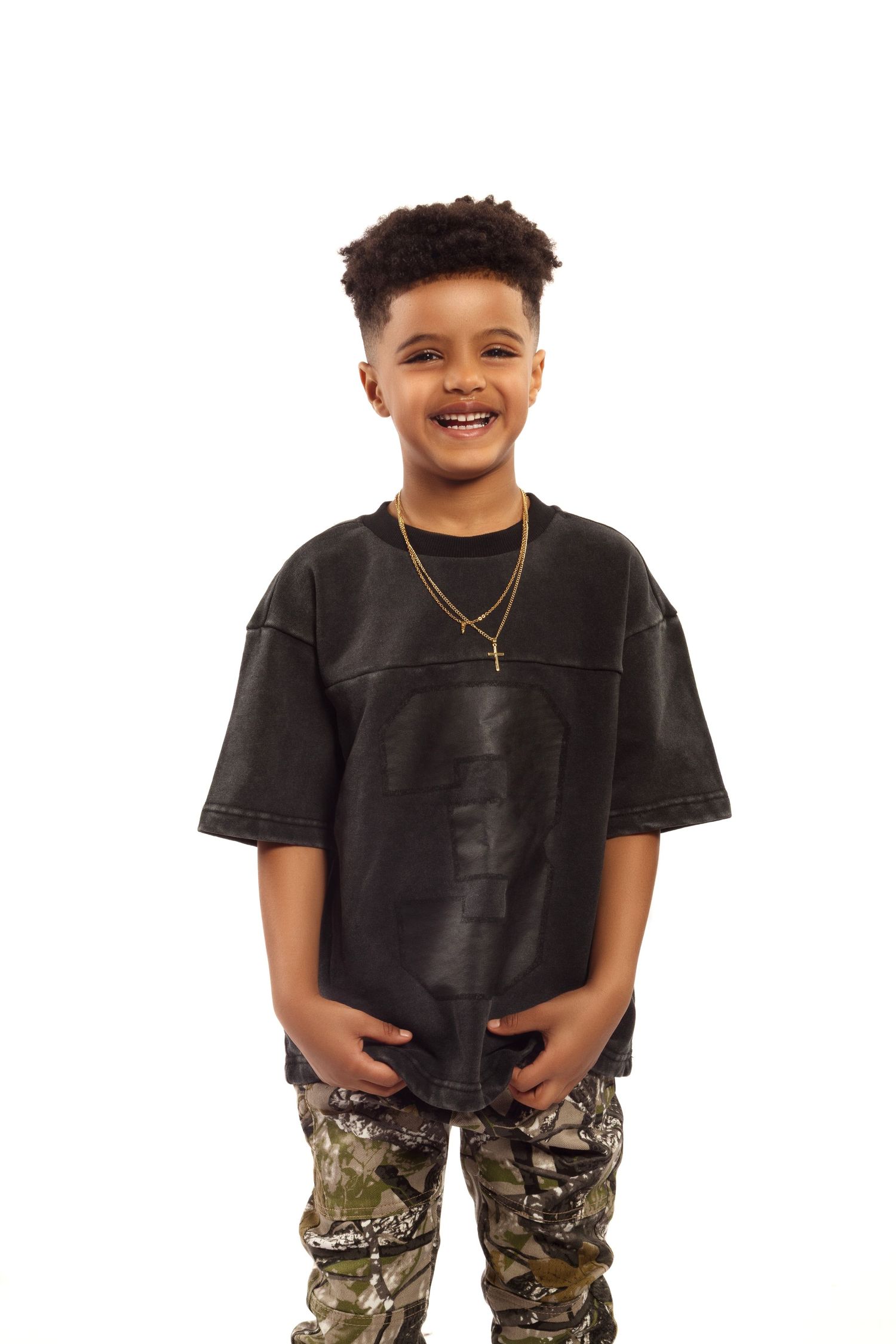 Young person wearing a black t-shirt and camouflage pants smiles while standing against white background.