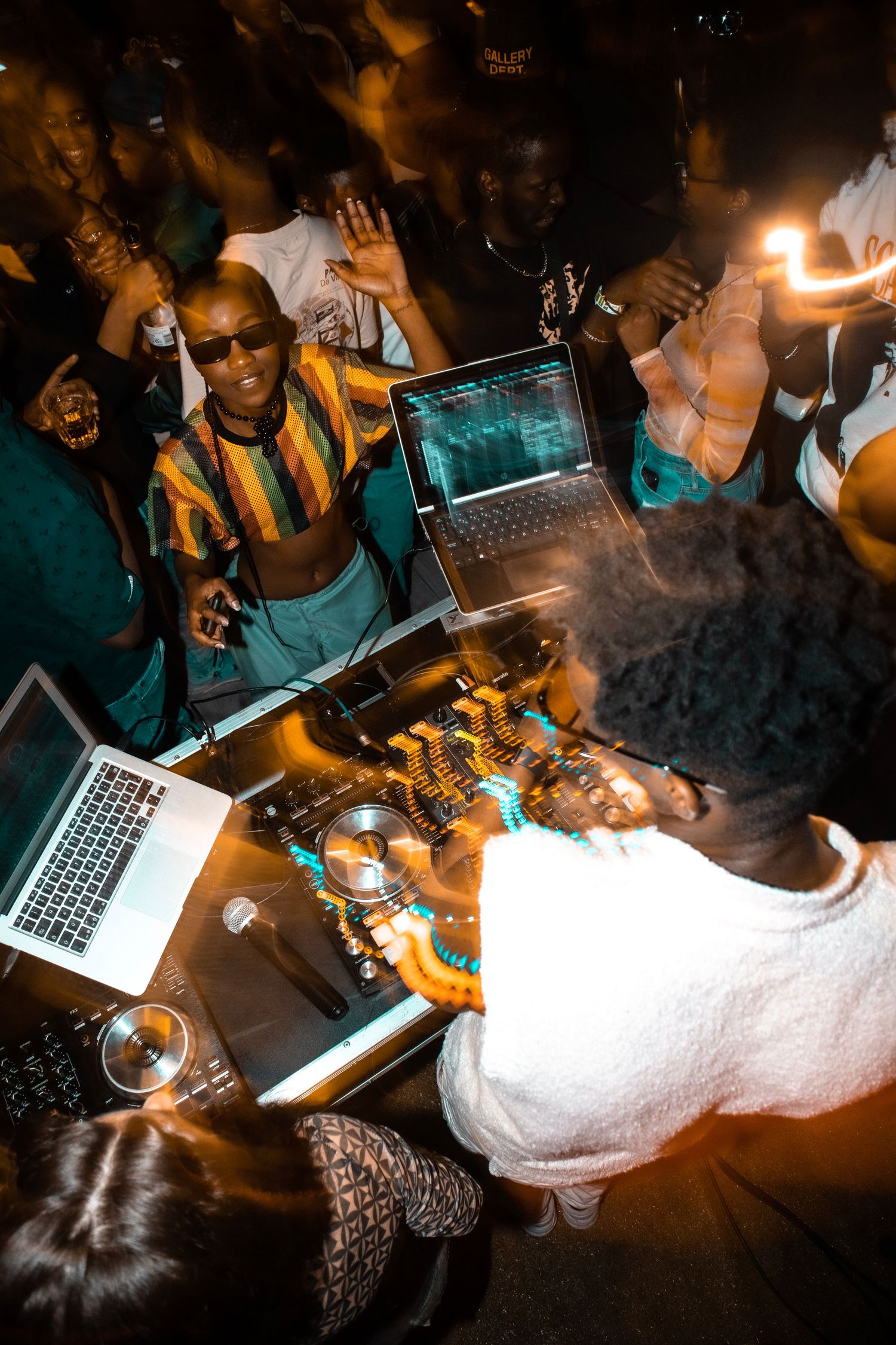 DJ setup at a crowded nightclub with laptops and mixing equipment illuminated by warm lighting.