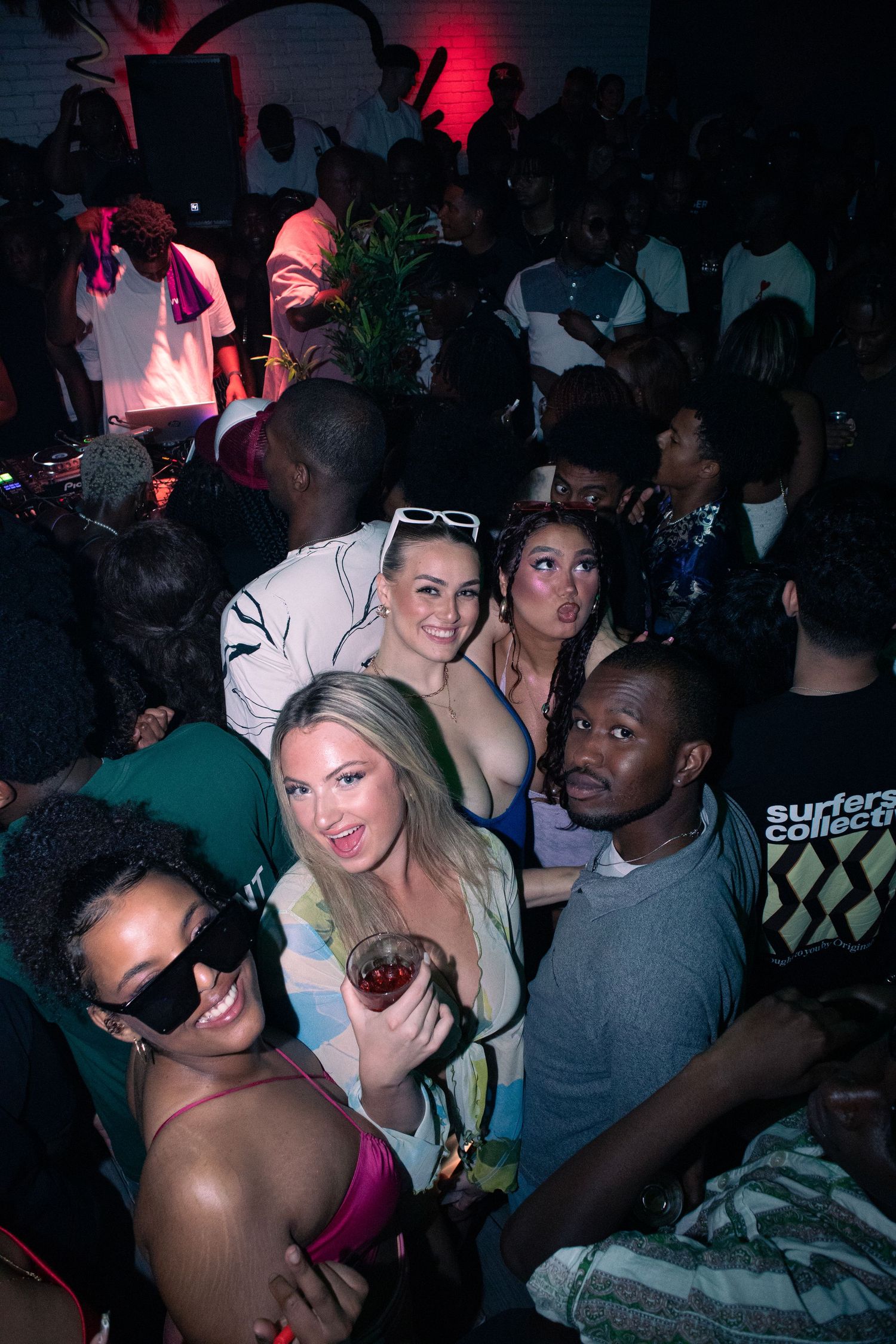 Crowded nightclub scene with partygoers dancing under red and purple lighting.