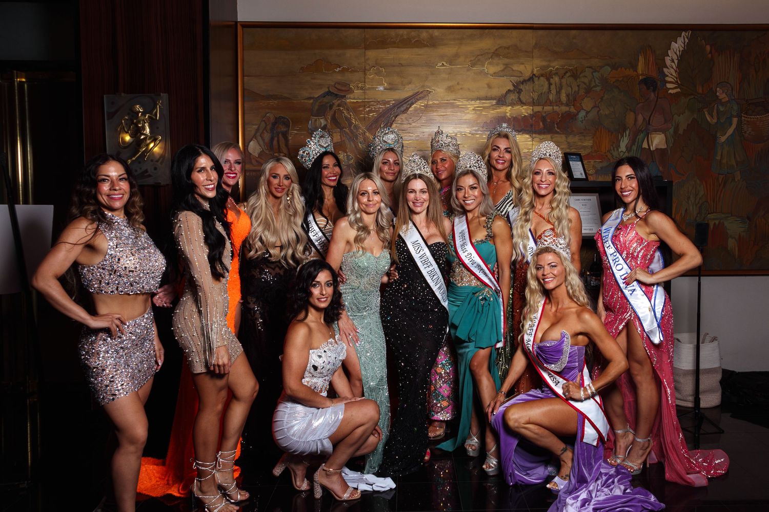 Group of women in elegant gowns and sashes posing together at a pageant event in a decorated venue.