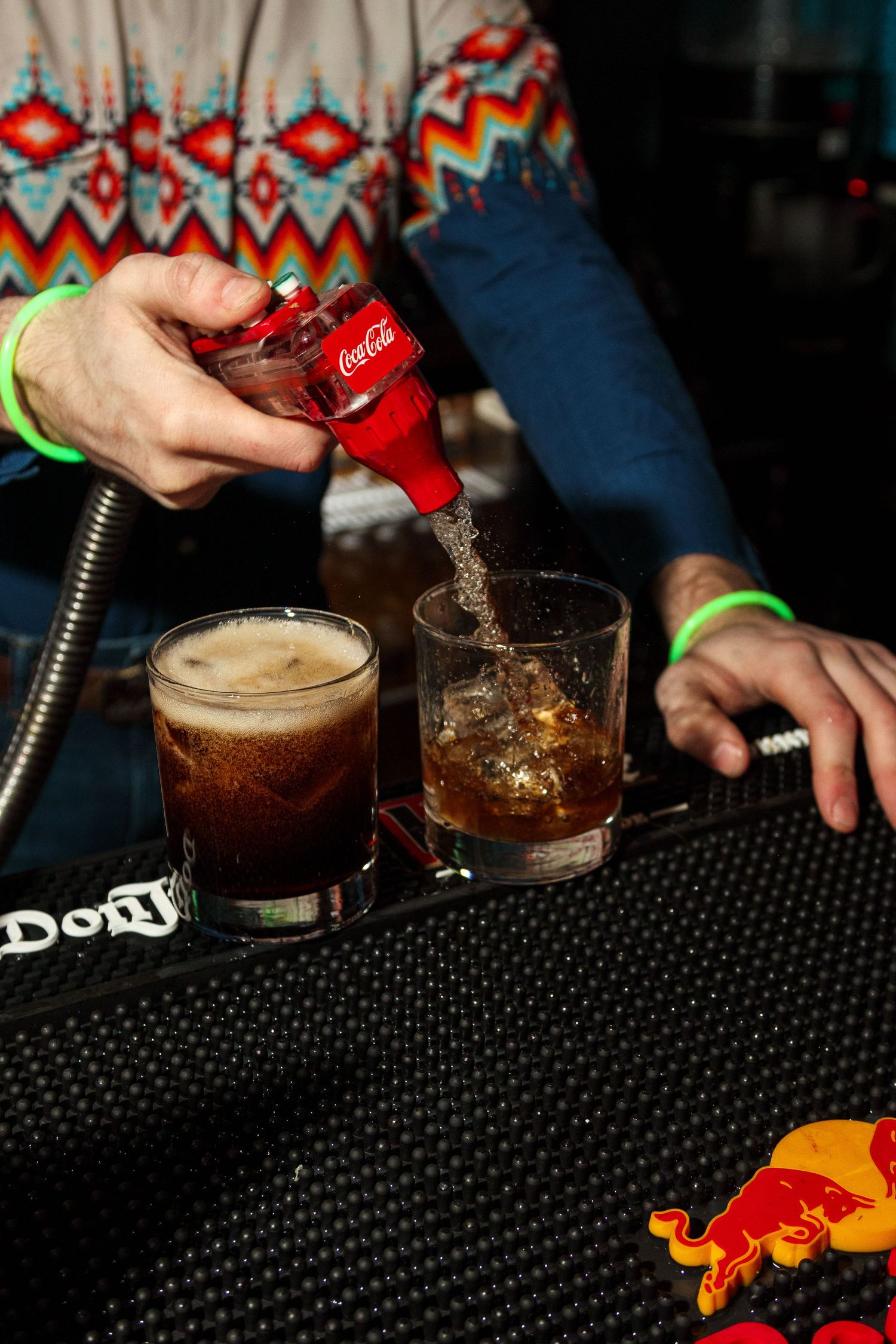 Bartender pouring Coca-Cola into mixed drinks at a bar with Red Bull branding visible.
