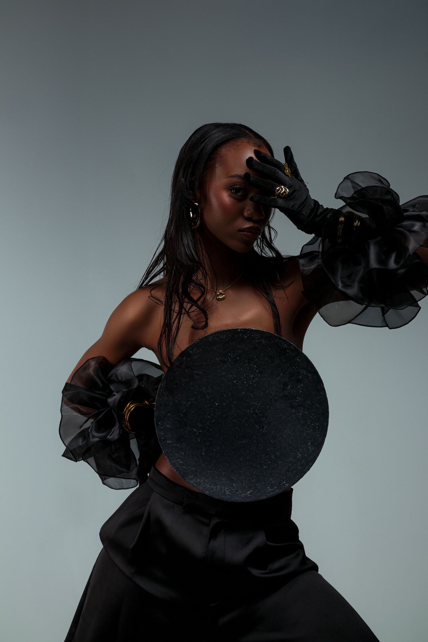 Dramatic fashion portrait with person in black outfit holding circular prop against gray background.