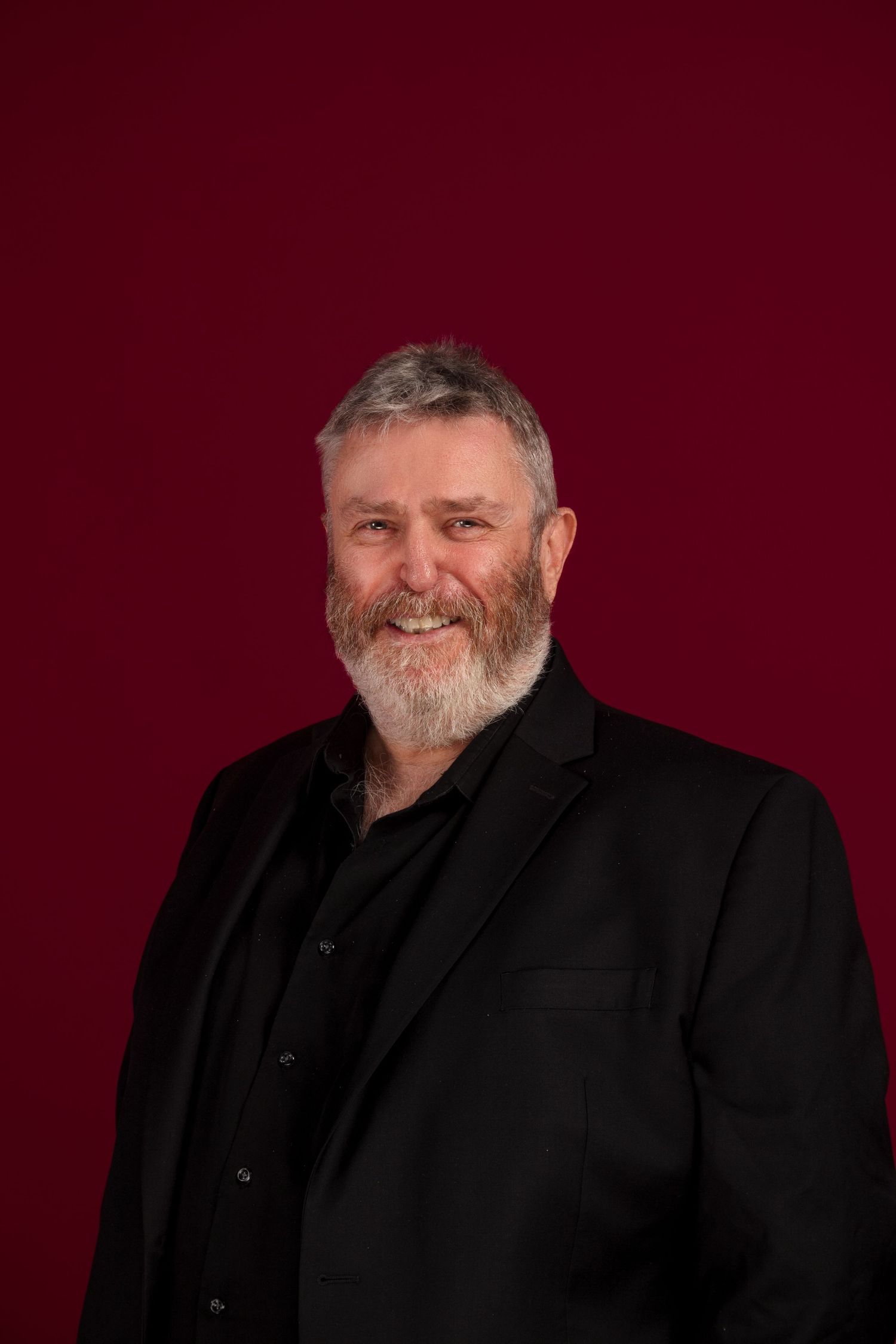 Professional headshot of bearded man in black suit against burgundy background.
