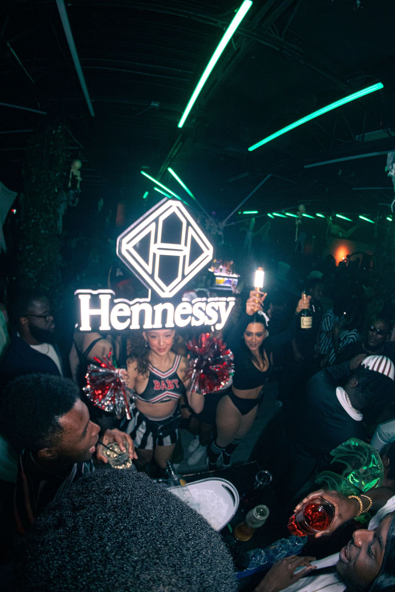 Neon Hennessy sign glowing in a dark nightclub with party crowd below.