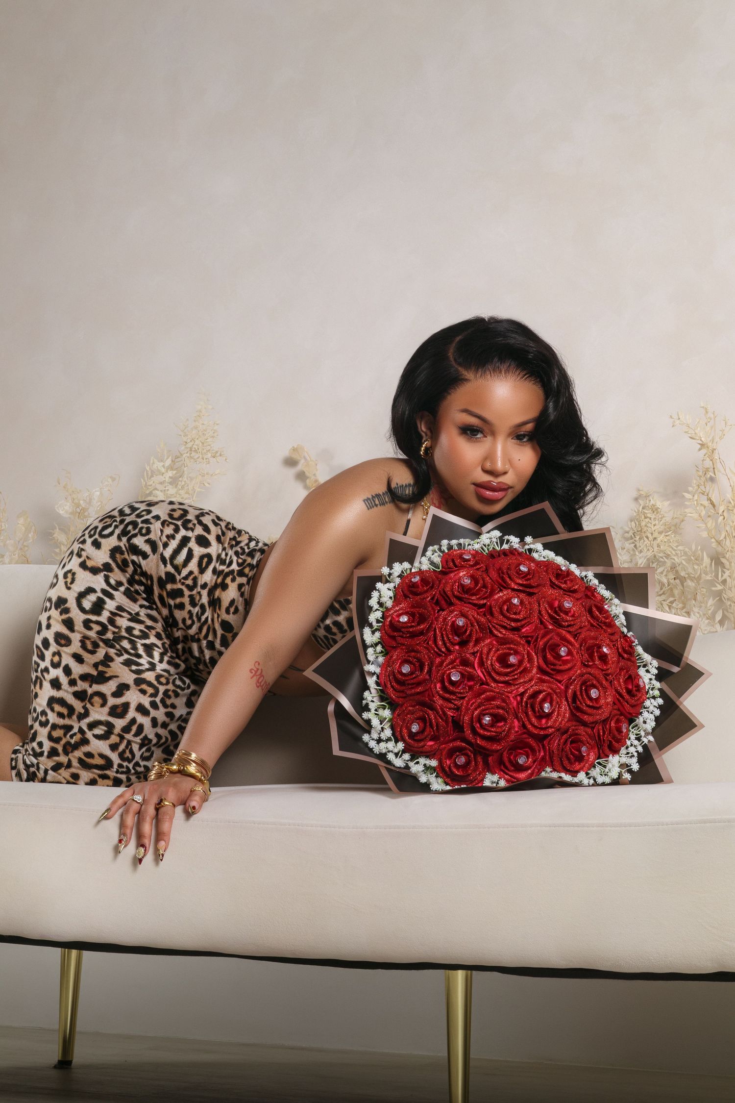 Glamorous photo of woman in leopard print dress posing on white couch with red rose bouquet.