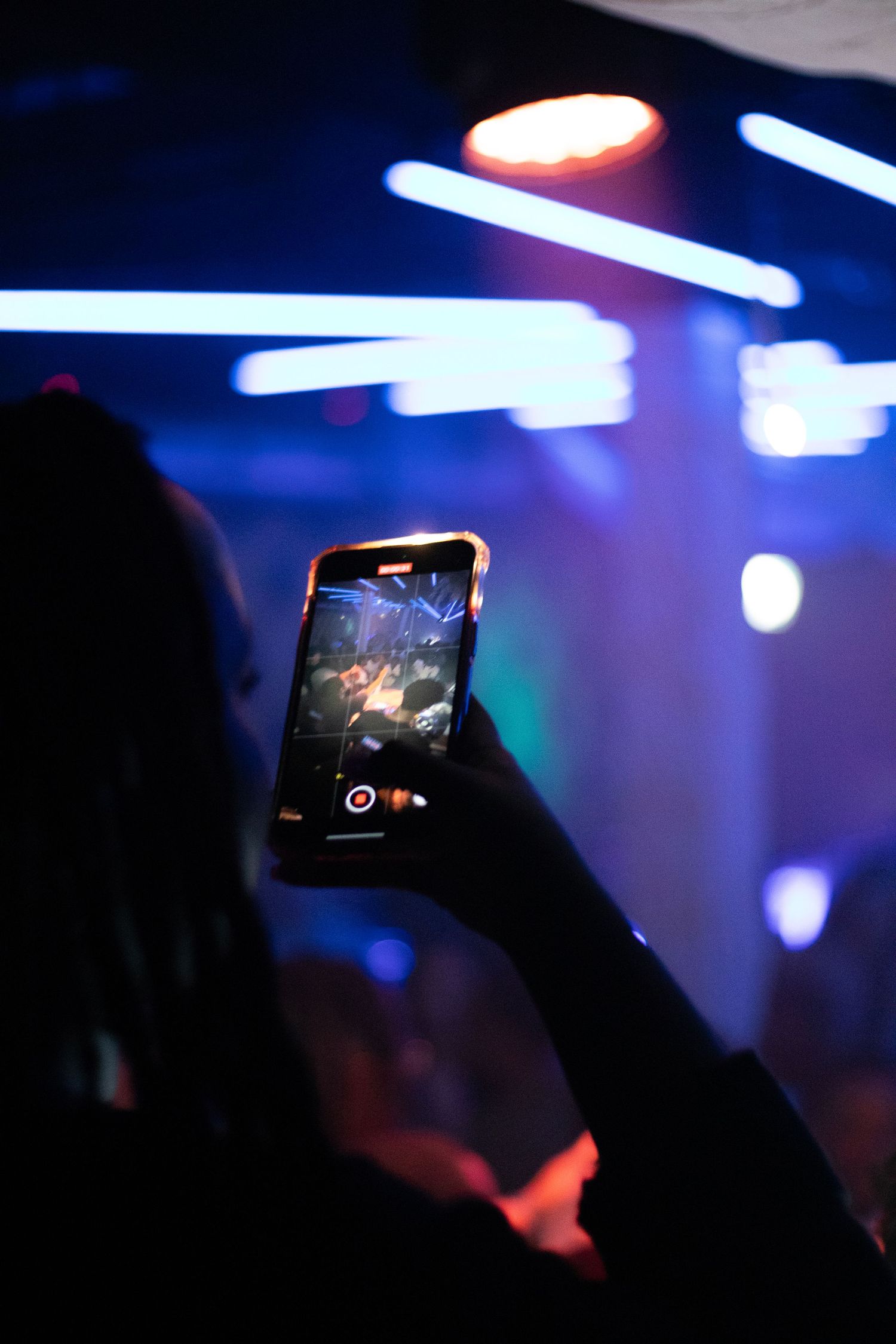 Silhouetted person captures concert footage on smartphone with blue stage lights in background.