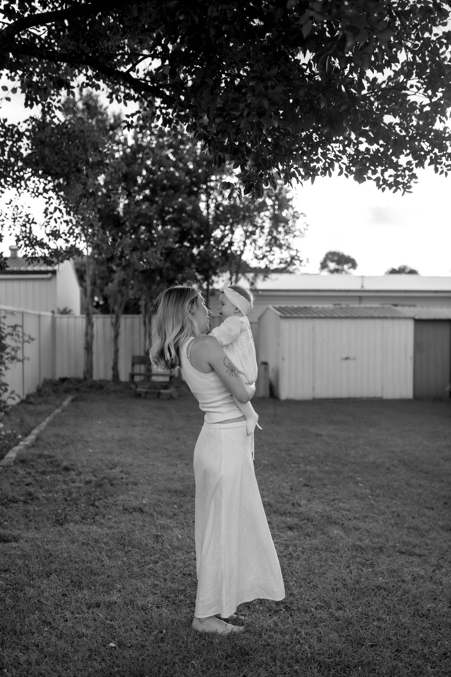 A tender black and white portrait of parent and baby embracing in a backyard garden setting.