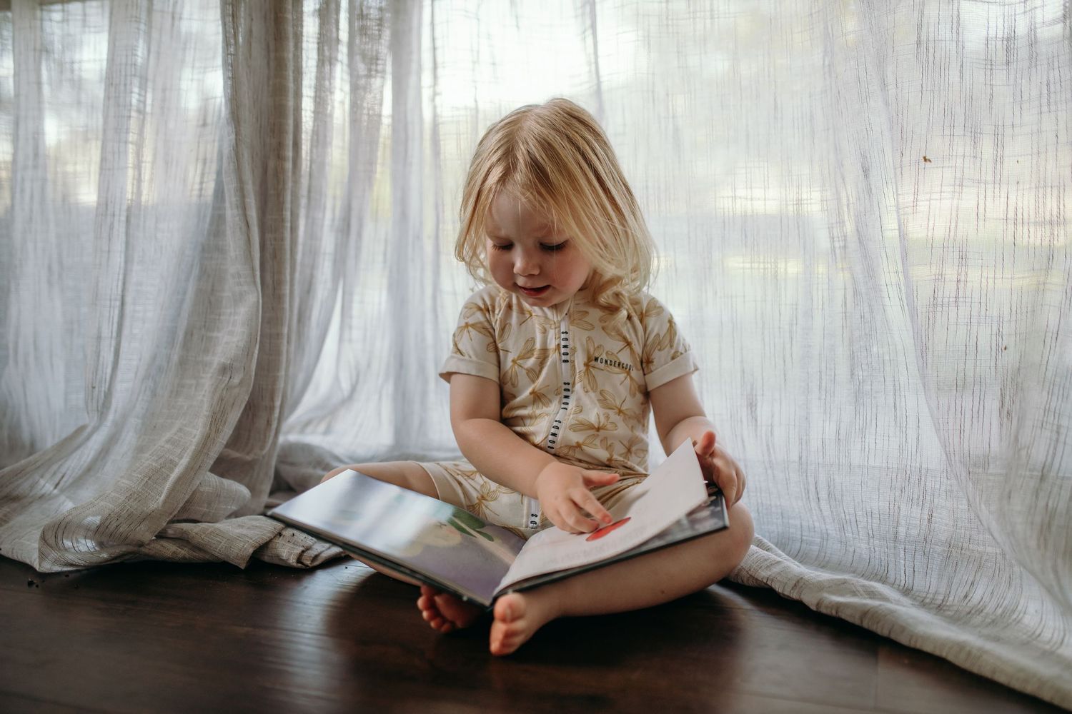 Child sitting by sheer curtains in natural light while reading.