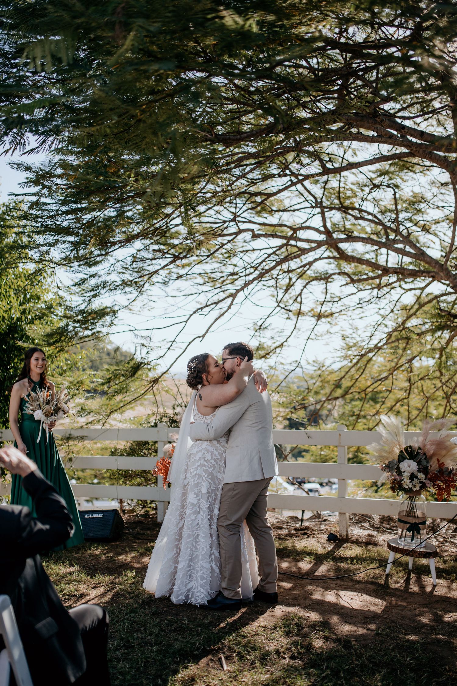 Outdoor wedding ceremony under large trees with guests watching the happy couple.