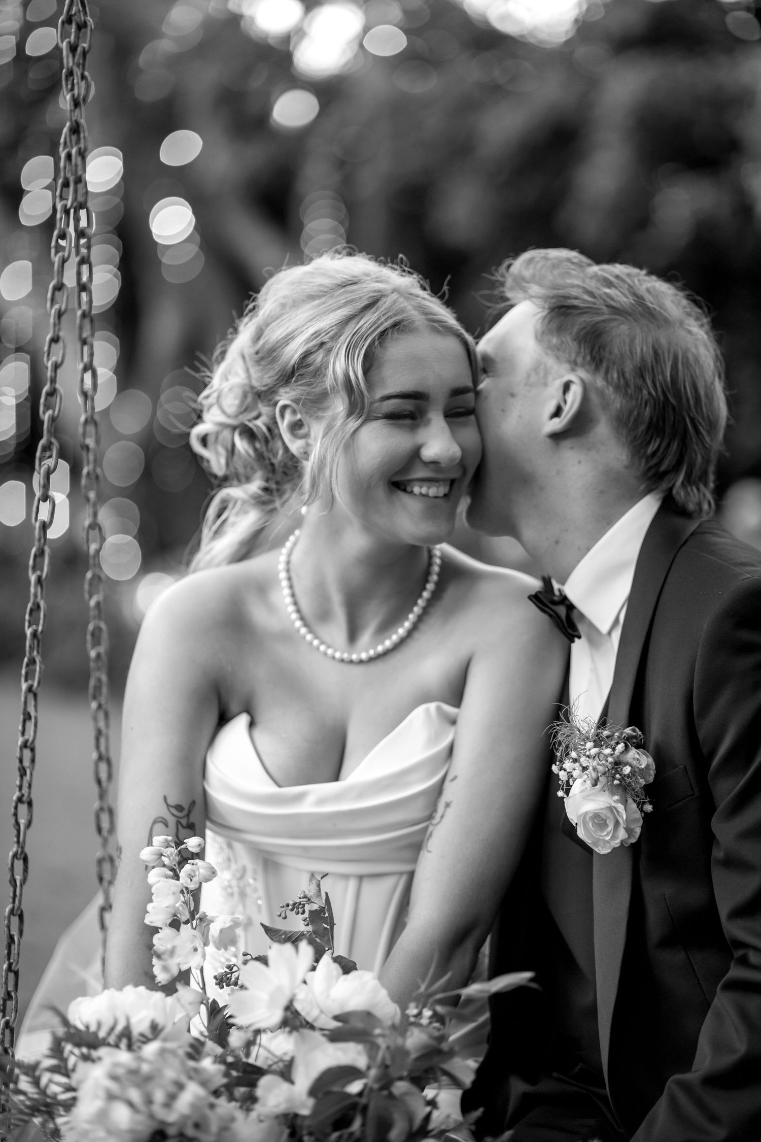 A joyful black and white wedding moment captured on a swing with bokeh lights in the background and floral bouquet details.