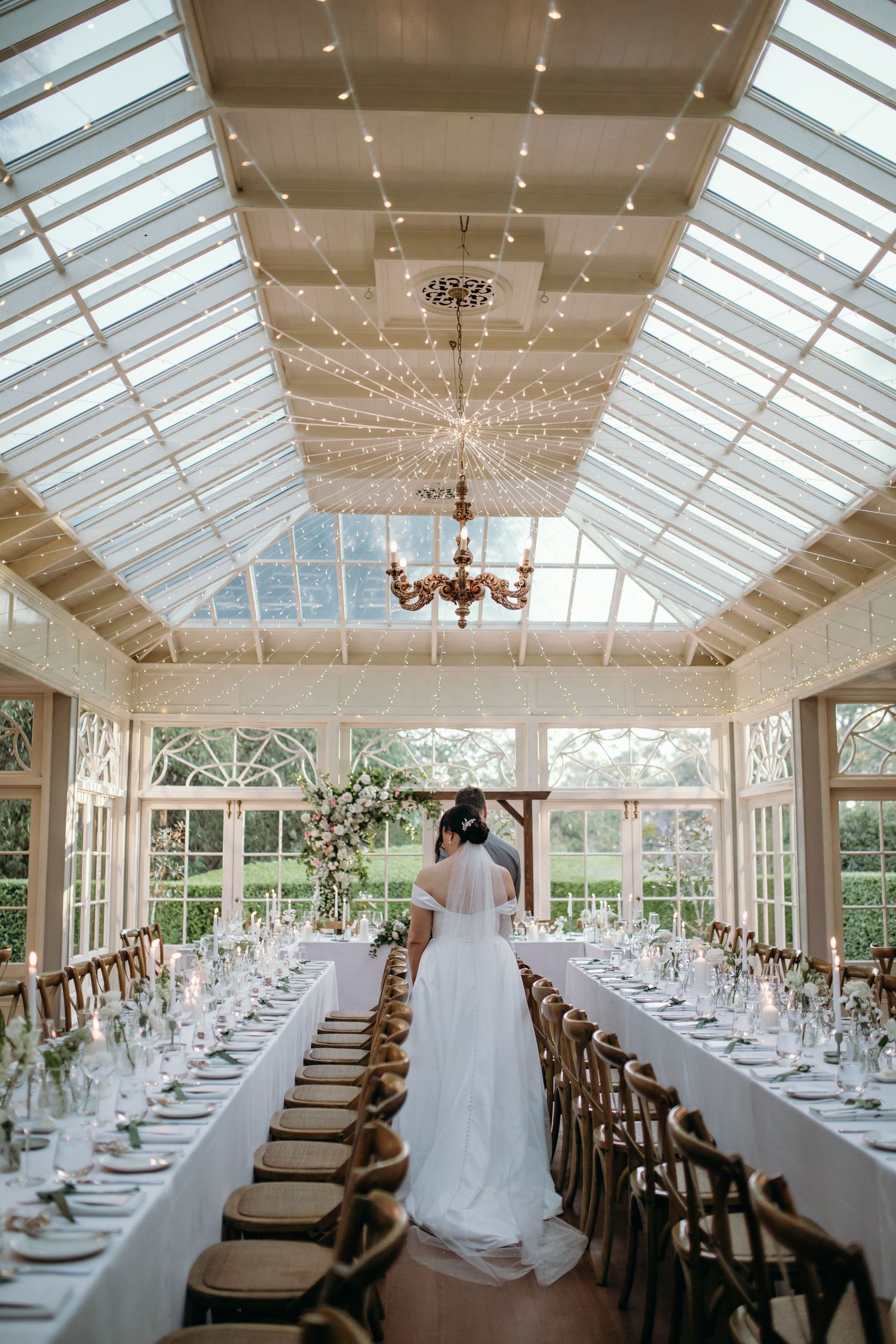 Elegant conservatory wedding reception setup with long white-dressed tables under a glass ceiling with crystal chandeliers.