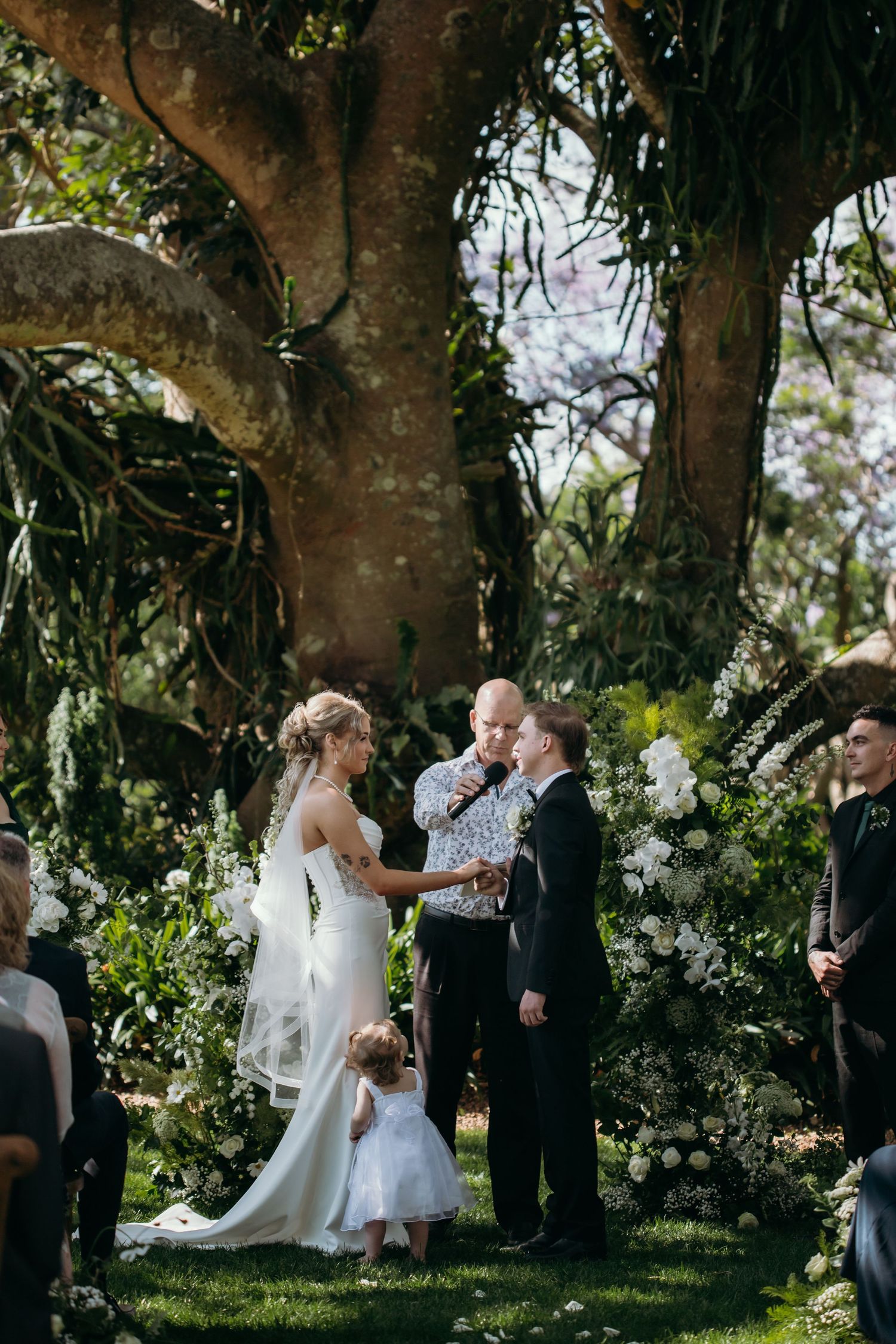 Wedding ceremony taking place under large trees decorated with white flowers.