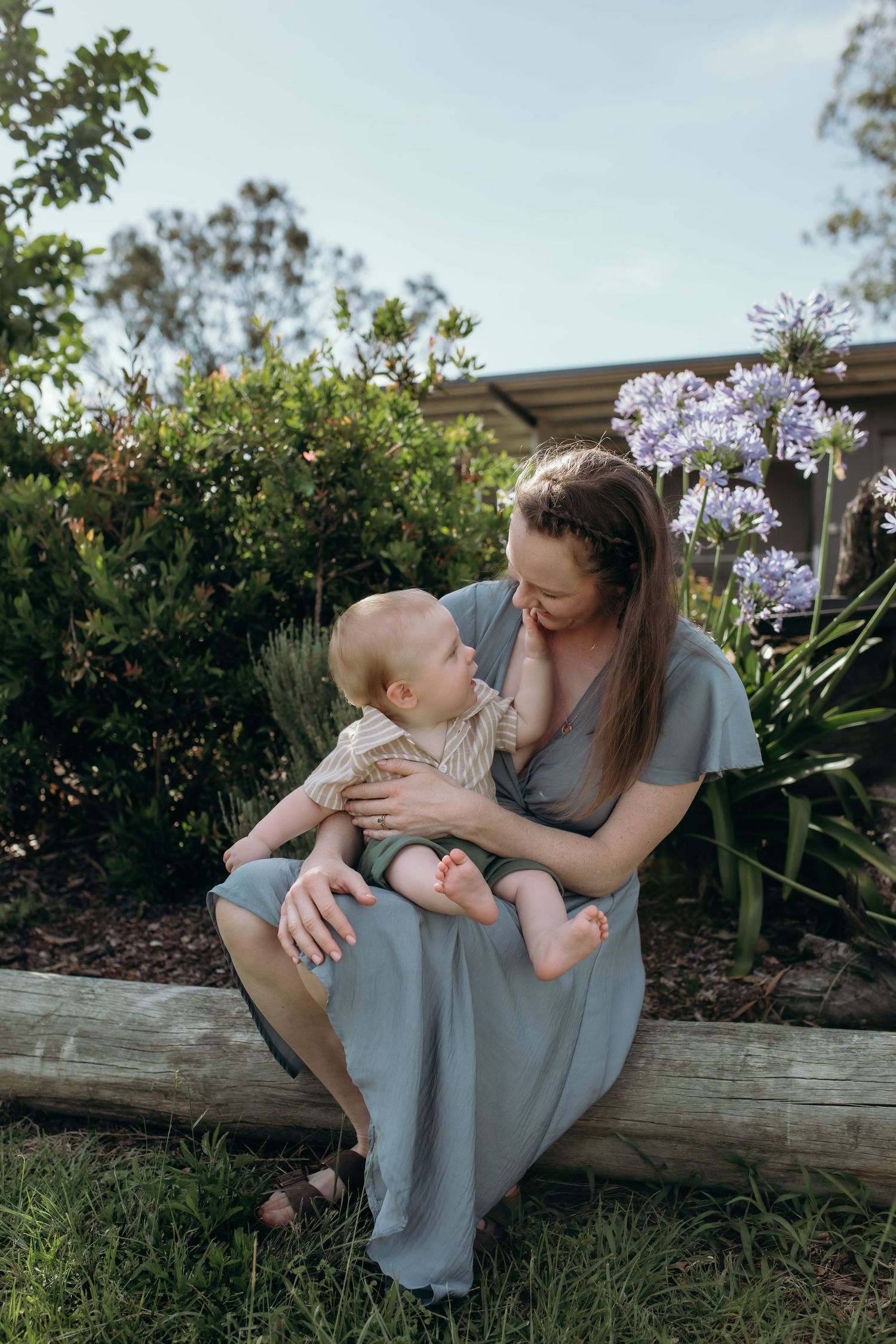Mother and baby share tender moment on log near blooming agapanthus flowers.