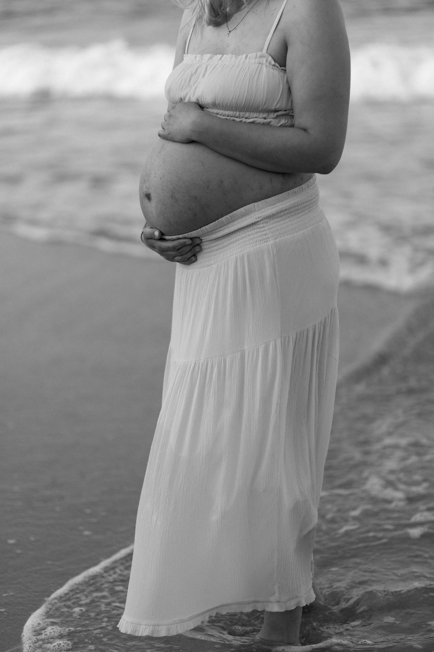 Silhouette of a pregnant figure in flowing white dress standing in ocean waves.