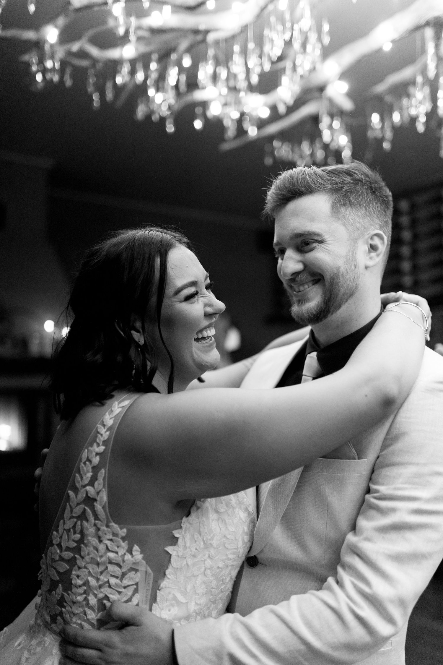 Black and white portrait of couple laughing together under crystal chandeliers.