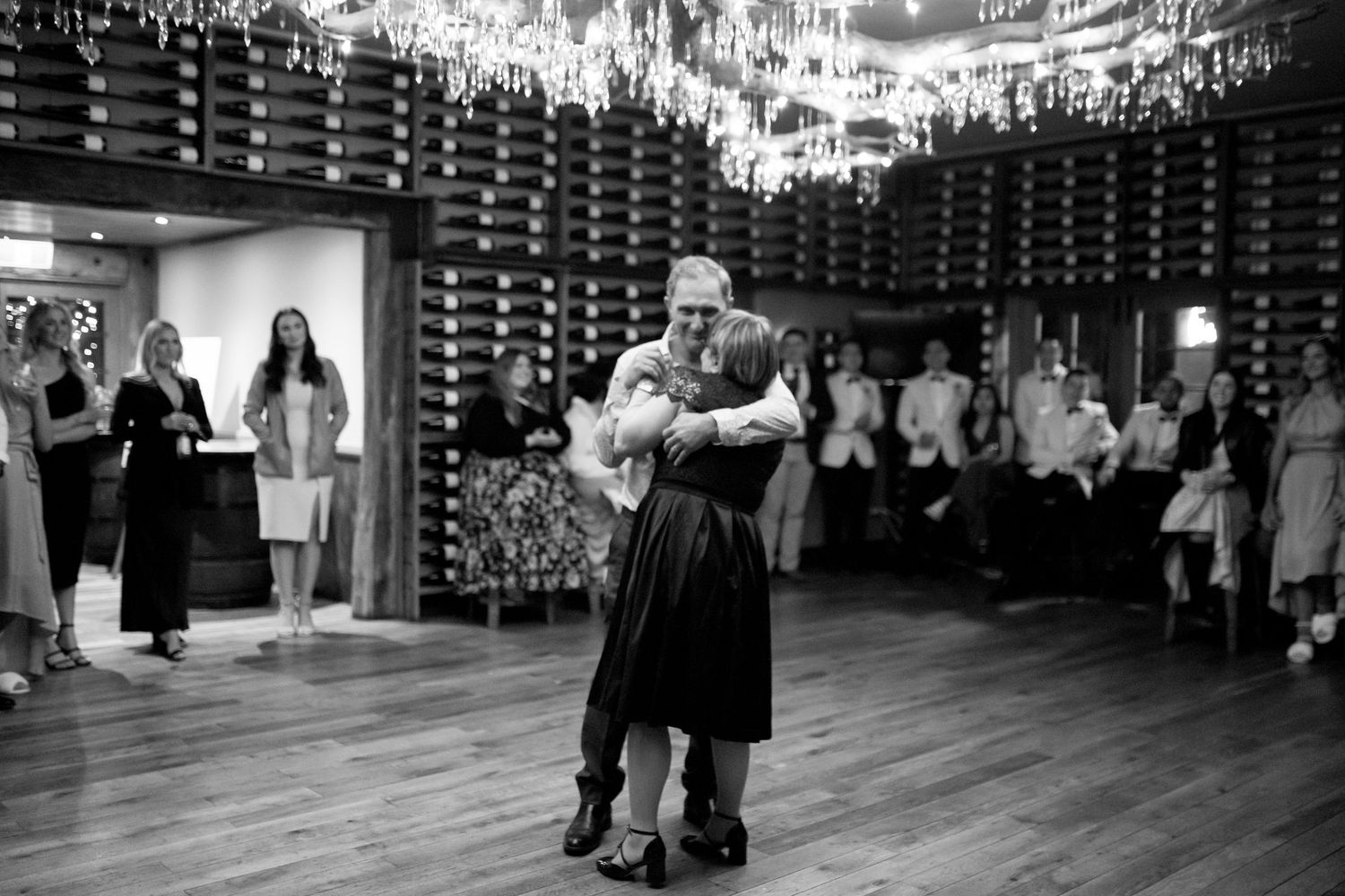 Black and white photo of couple dancing in elegant indoor venue with chandeliers.