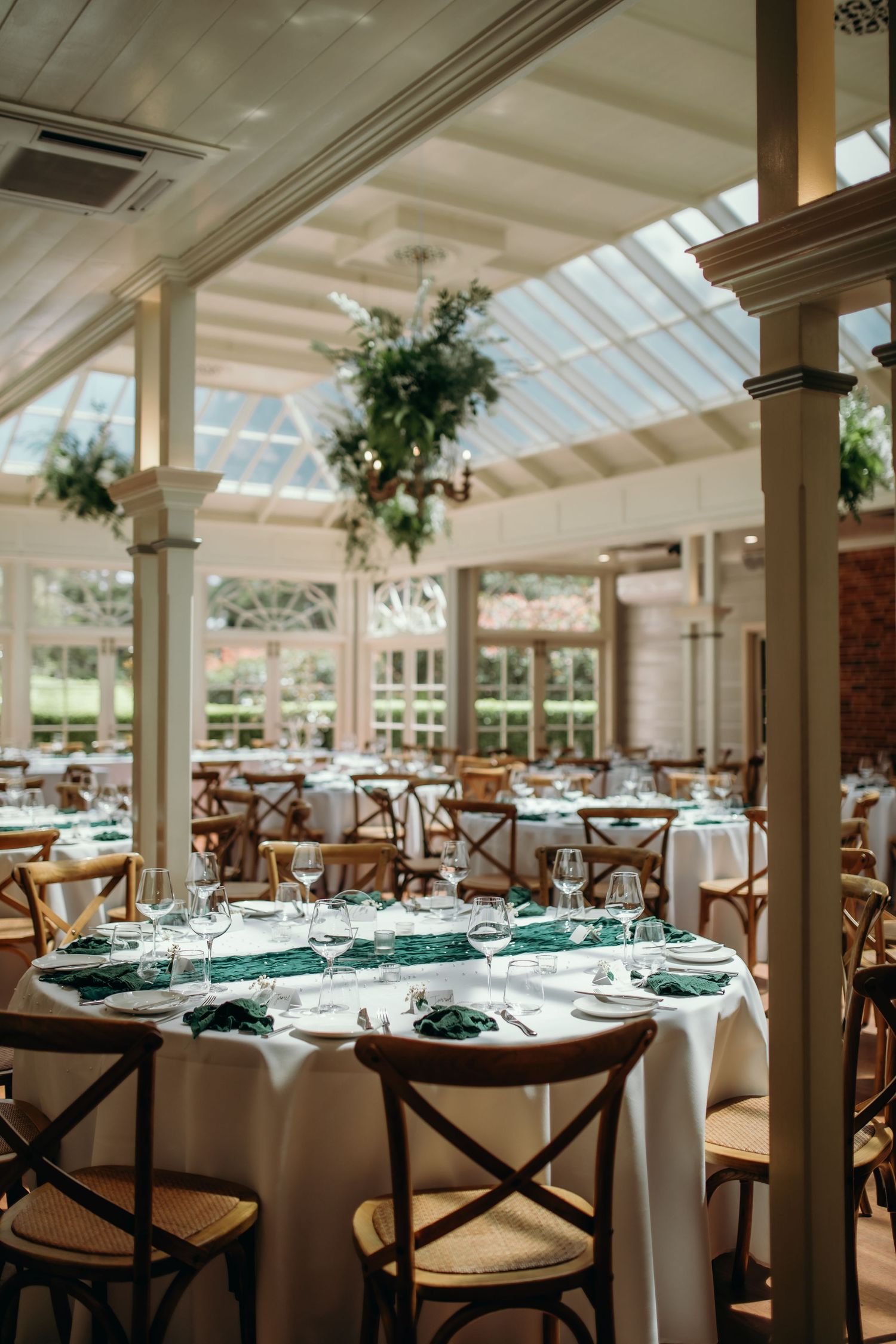 Elegant dining room setup with white tablecloths, teal napkins, and cross-back chairs under a bright conservatory ceiling.