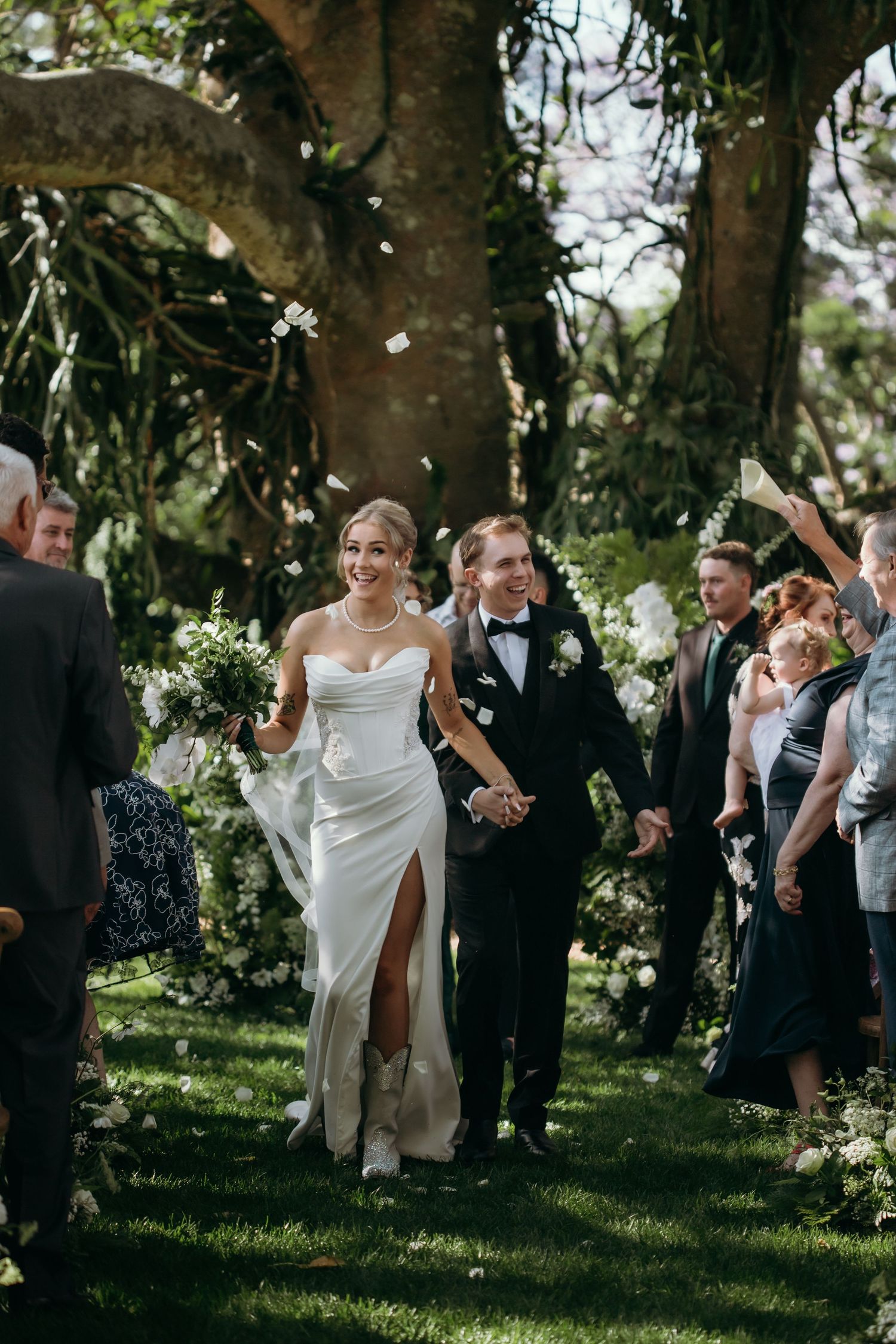 Wedding celebration with guests throwing confetti under tree canopy.