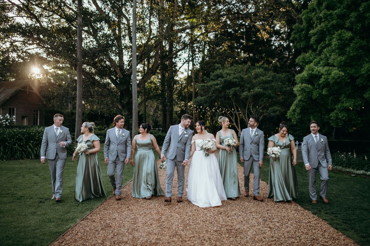 A wedding party in grey and sage green attire poses together on a tree-lined path.