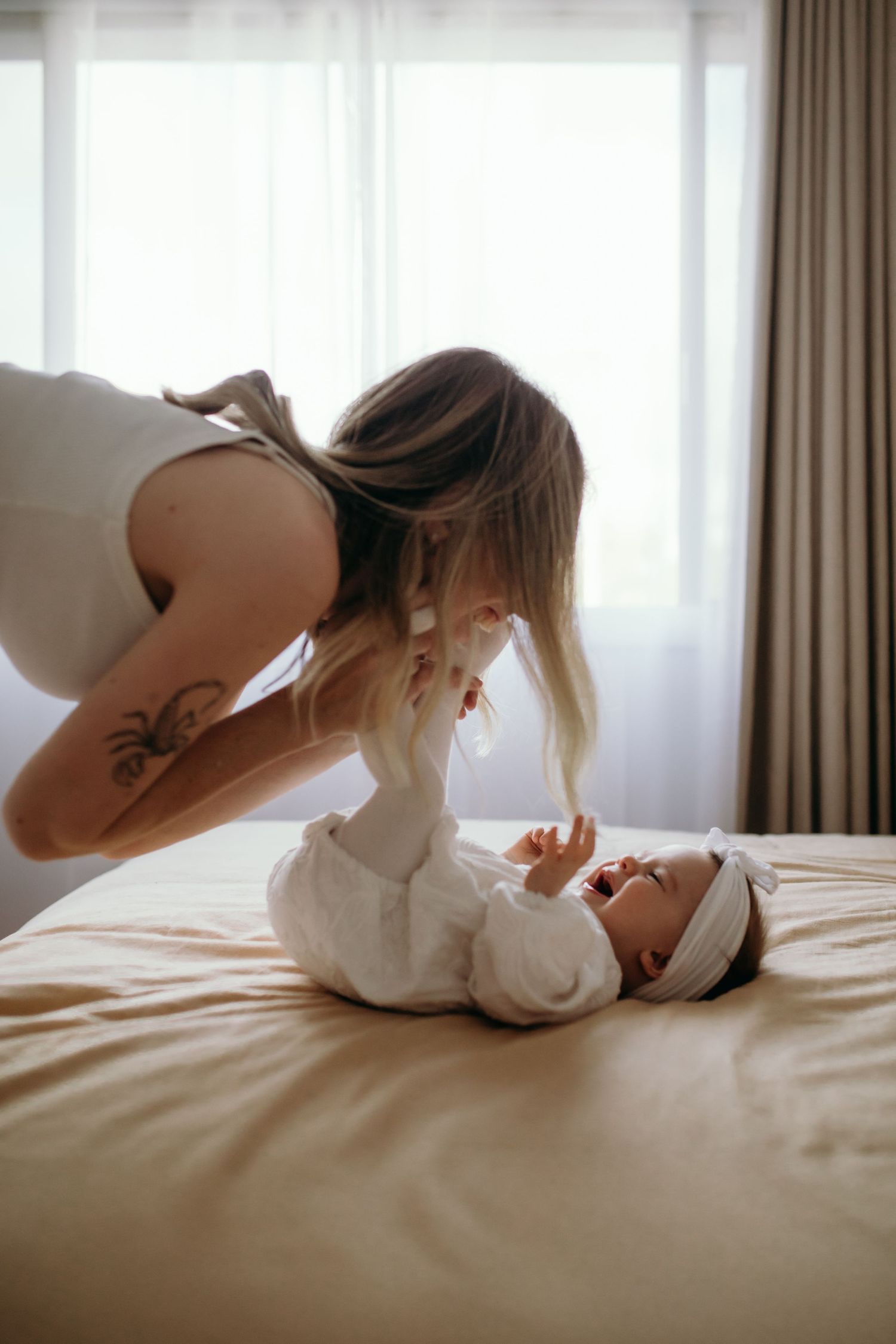 Parent playing with baby lying on bed in soft natural window light.
