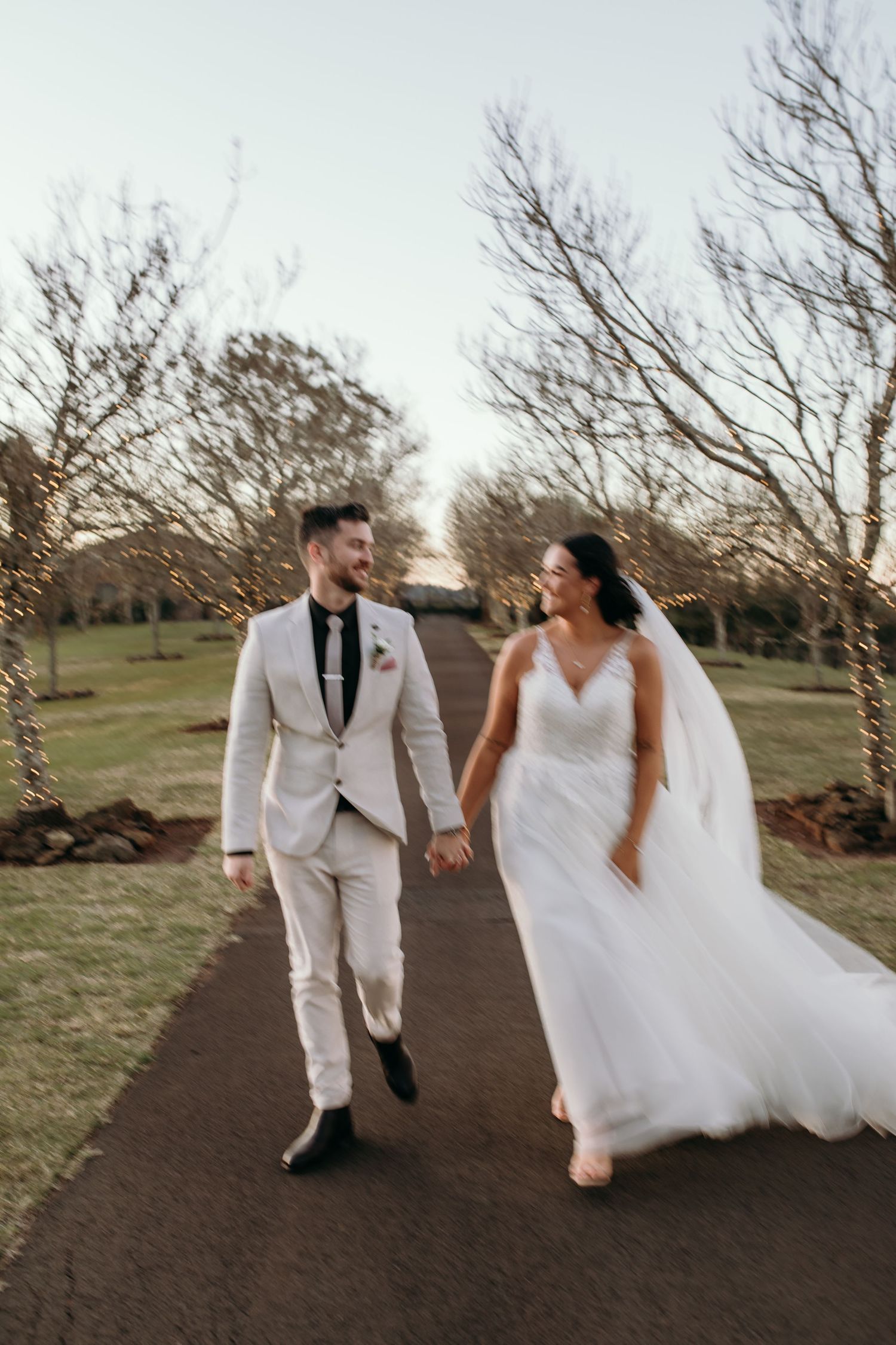 Newlyweds walk hand in hand along a winding path surrounded by bare trees.