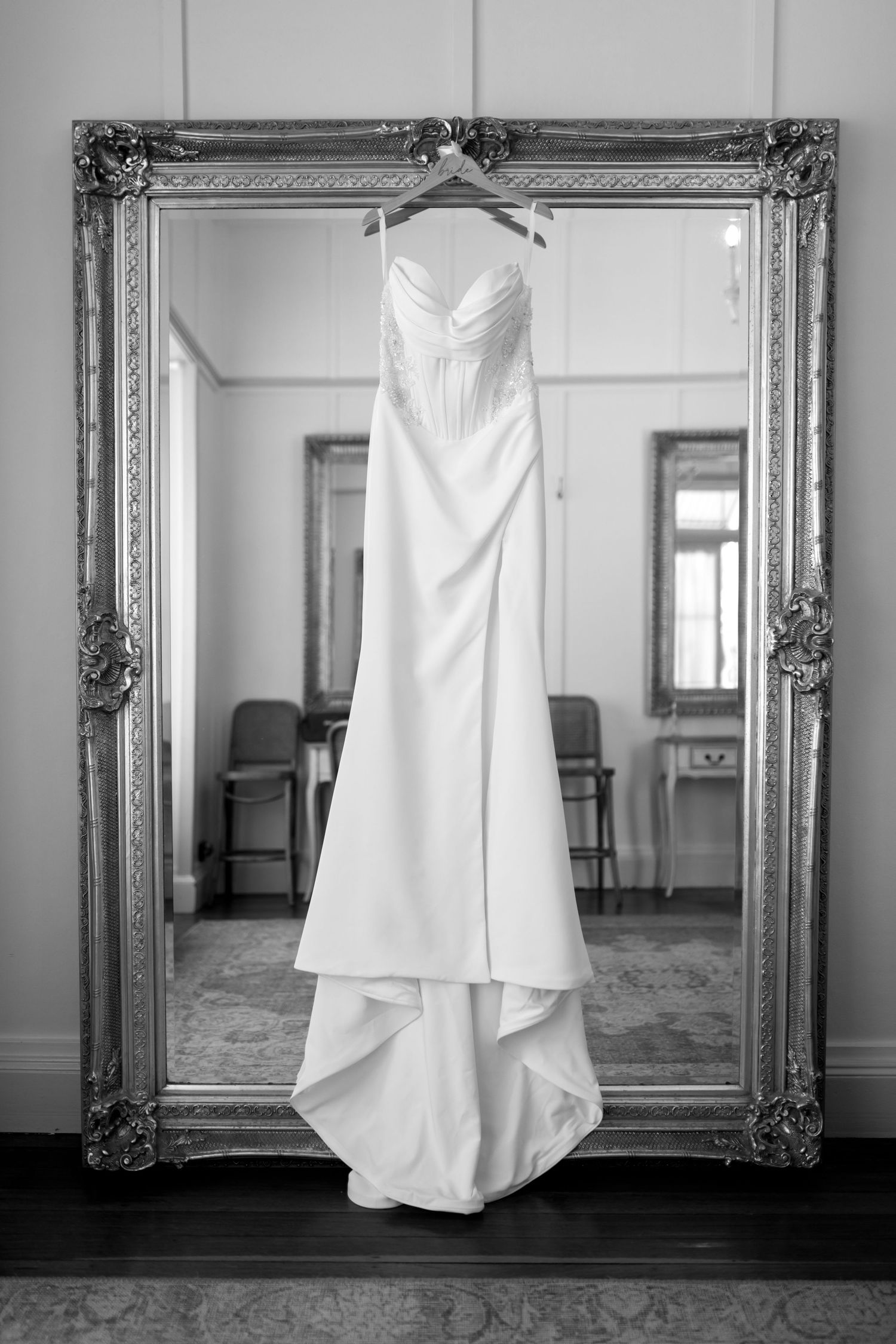 A white wedding dress hangs elegantly on a hanger in front of an ornate silver-framed mirror in a classic interior.