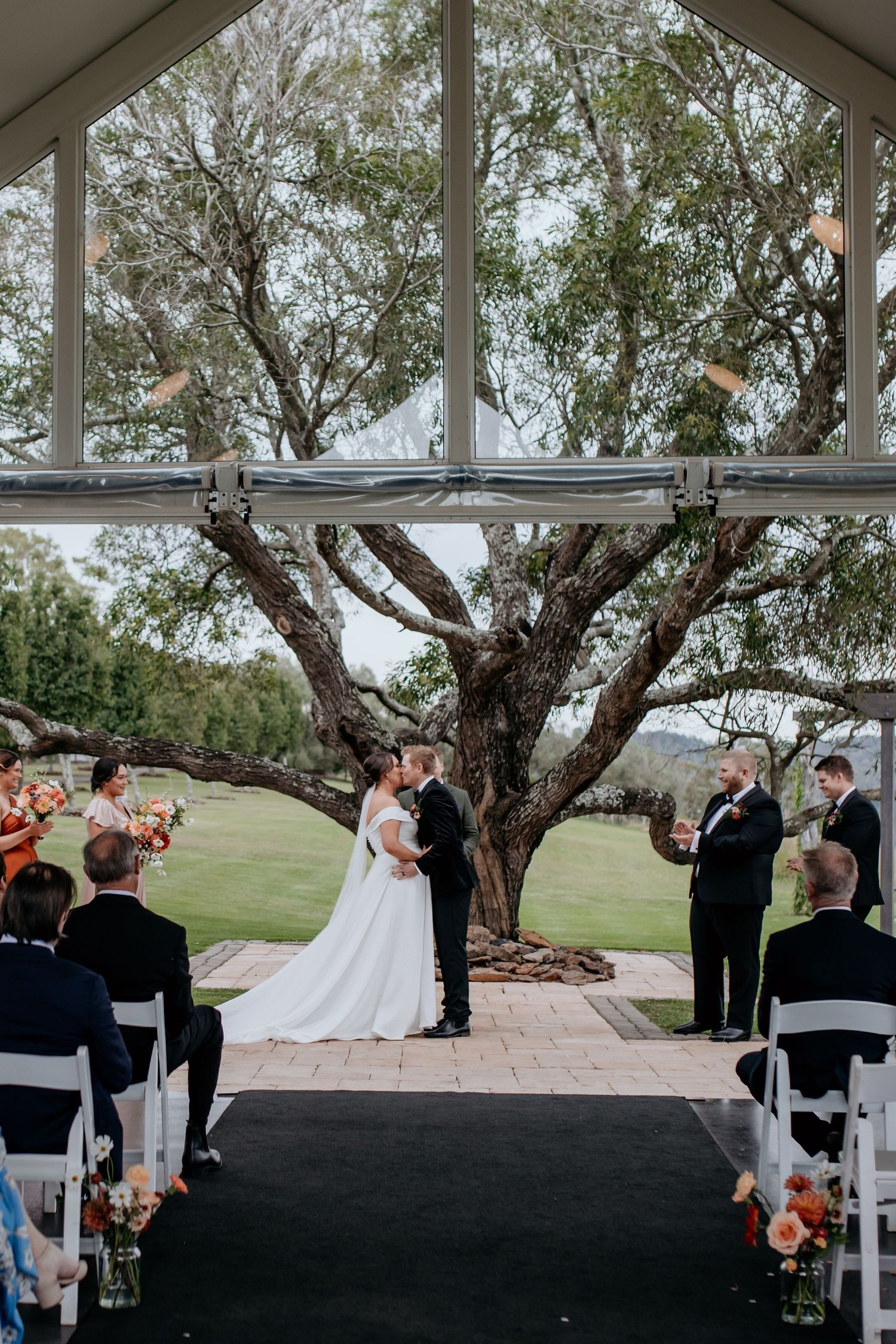 Panoramic view of an outdoor wedding ceremony under a white pavilion with guests seated and a large tree in the background.