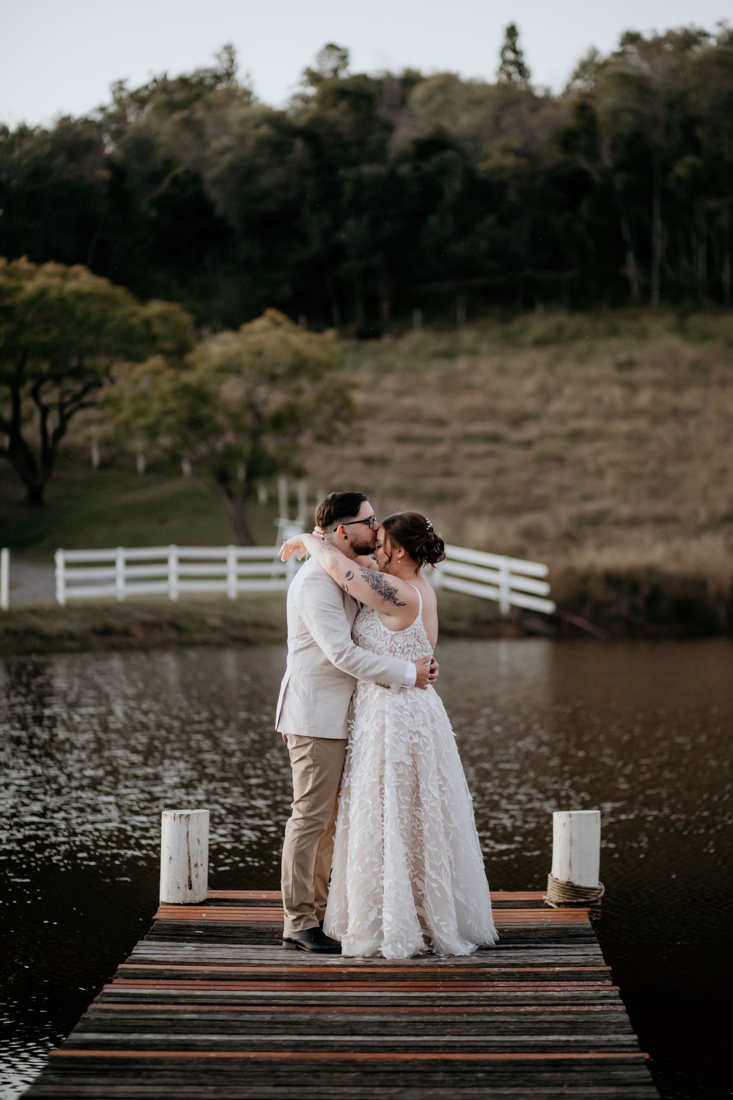 A romantic moment between newlyweds embracing on a wooden dock by a serene lake at sunset.