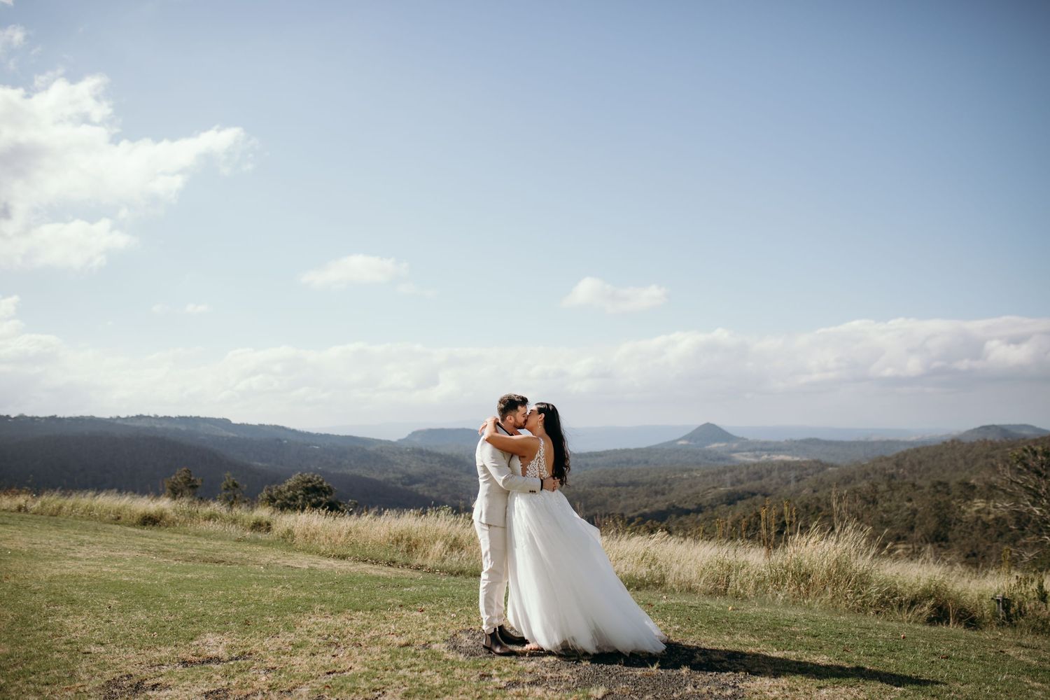 Newlyweds embrace on a hilltop with sweeping mountain views and dramatic sky in the background.