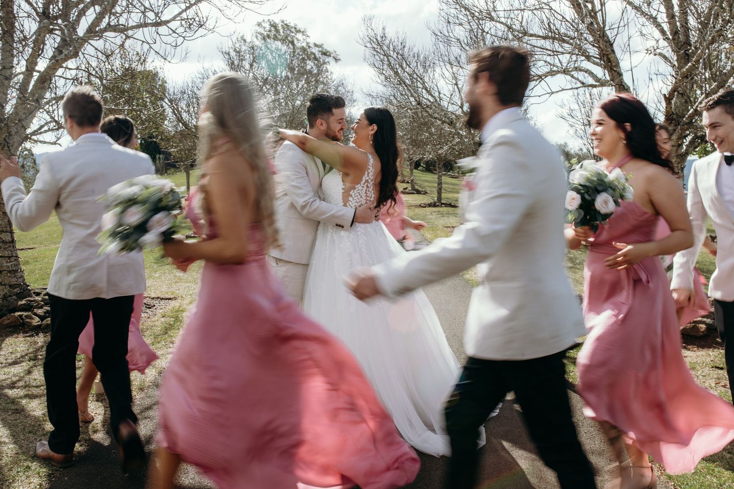 Wedding party dances joyfully outdoors with bridesmaids in flowing pink dresses.
