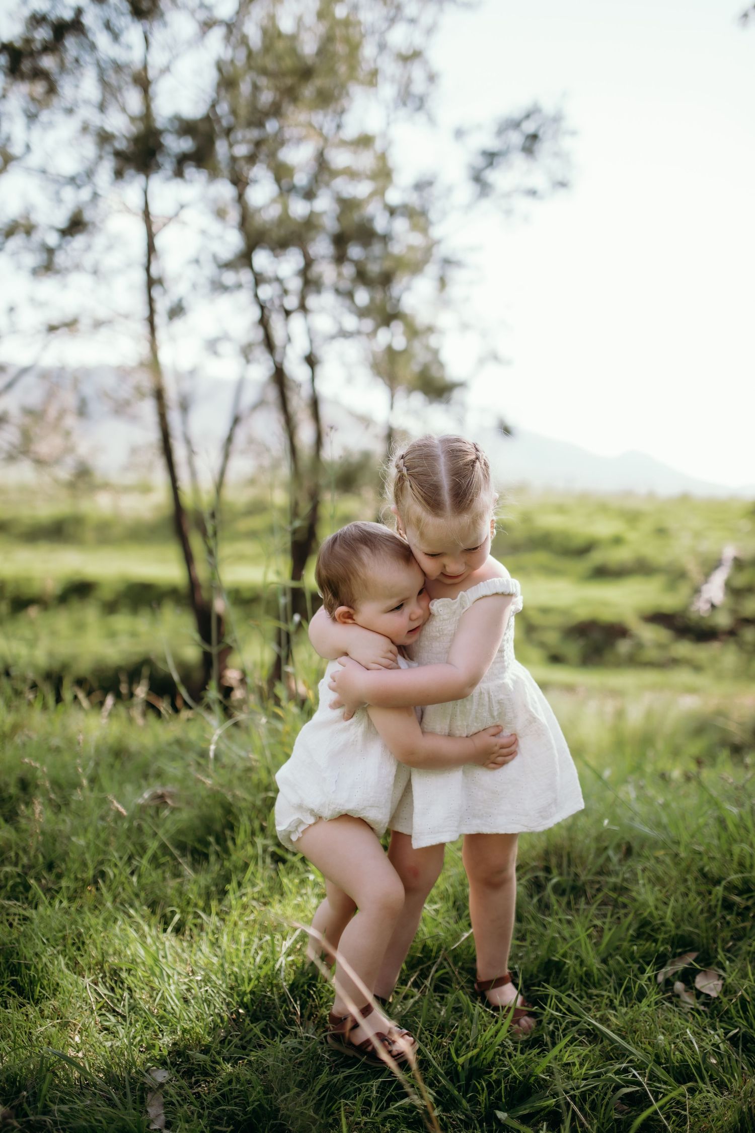 Two small children in white outfits sharing a sweet moment in a grassy field.
