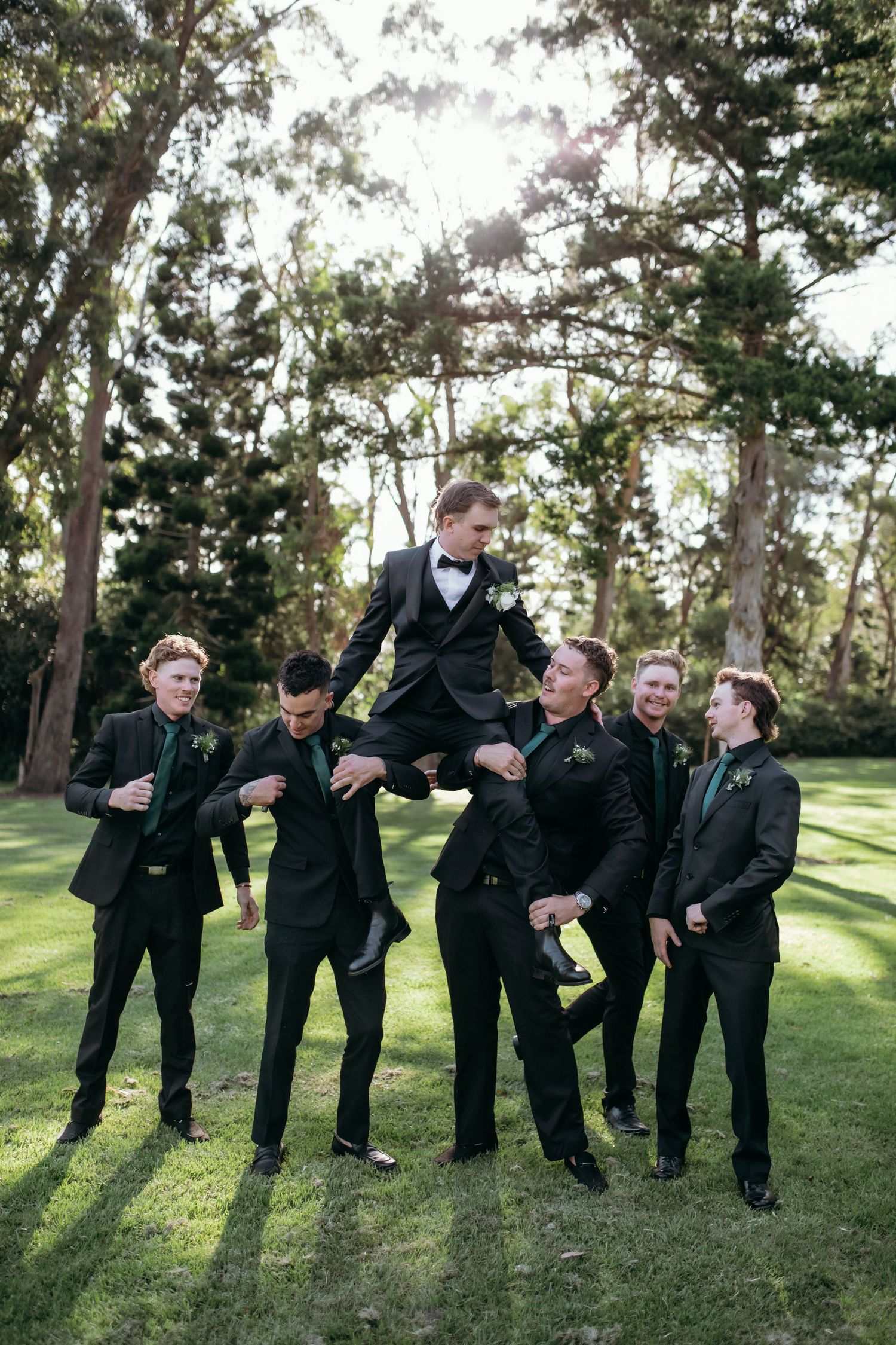 Groomsmen in black suits posing playfully on grassy lawn.