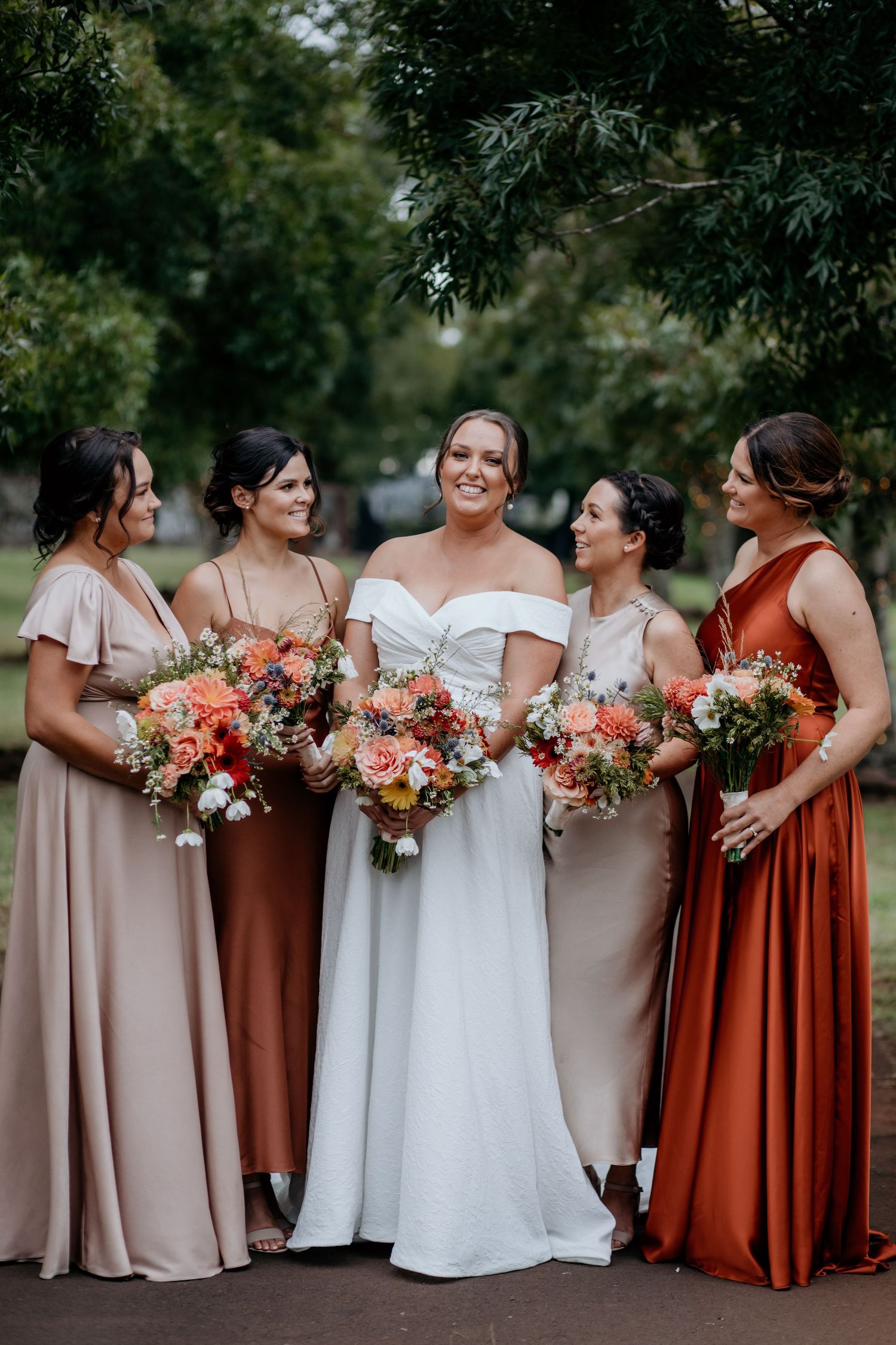 Wedding party in earth-toned dresses holding colorful floral bouquets stand together outdoors in a garden setting.