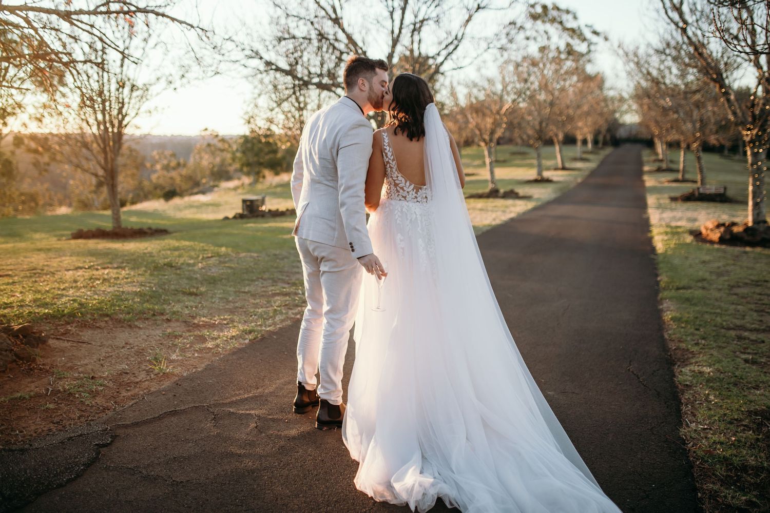 Bride and groom walk together on path surrounded by autumn trees at sunset.