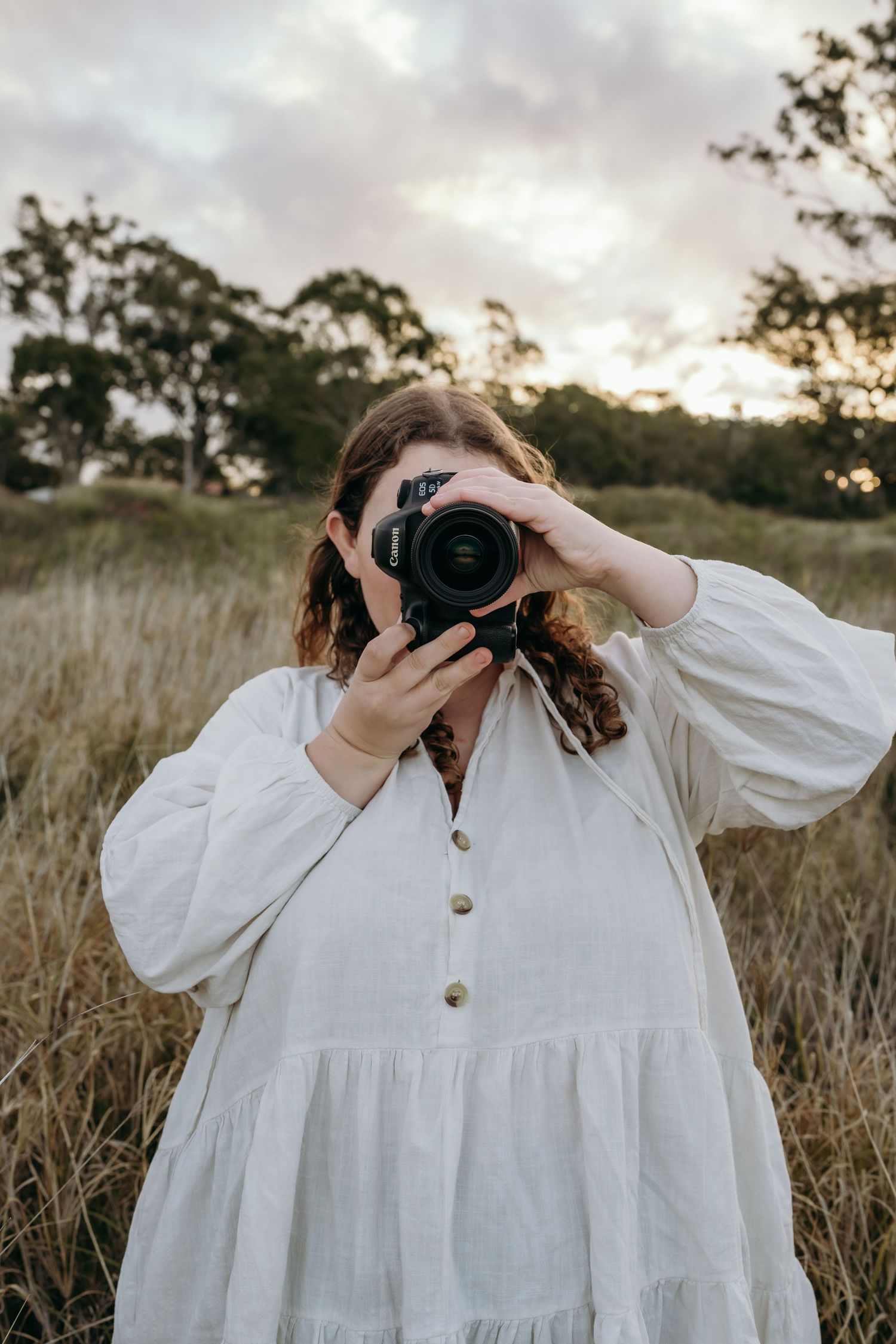 Person in flowing white dress taking photos with professional camera in field at sunset.
