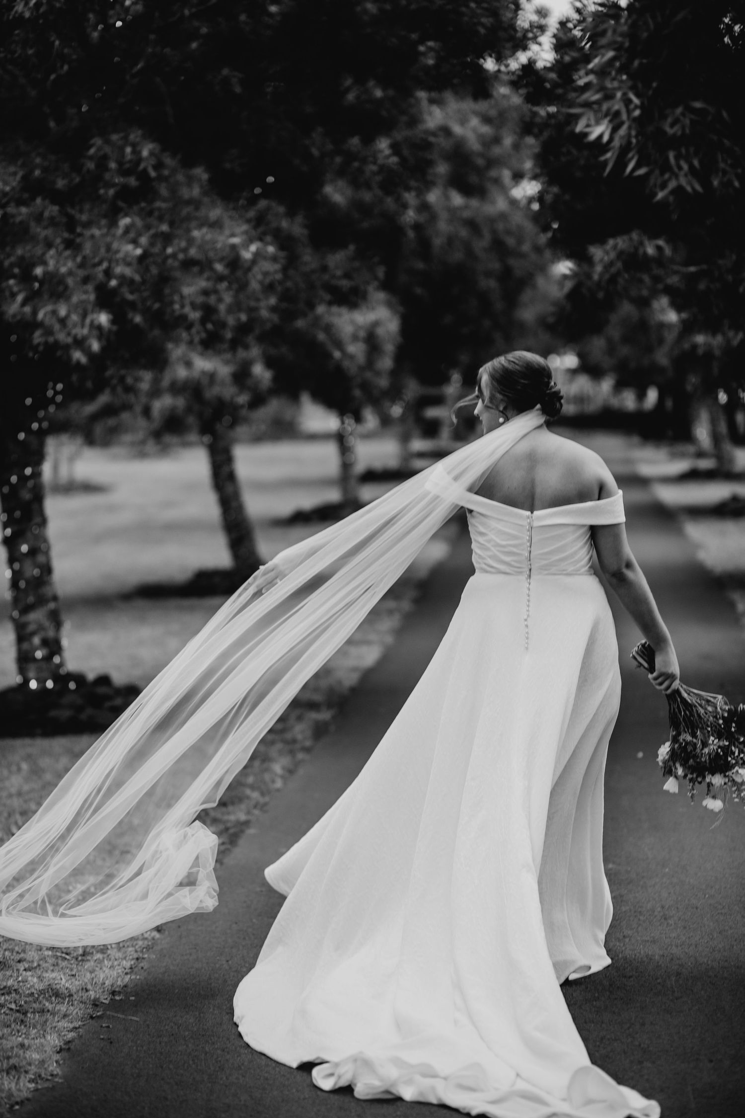 Black and white wedding photo showing a bride in an off-shoulder gown with flowing veil walking in a garden setting.