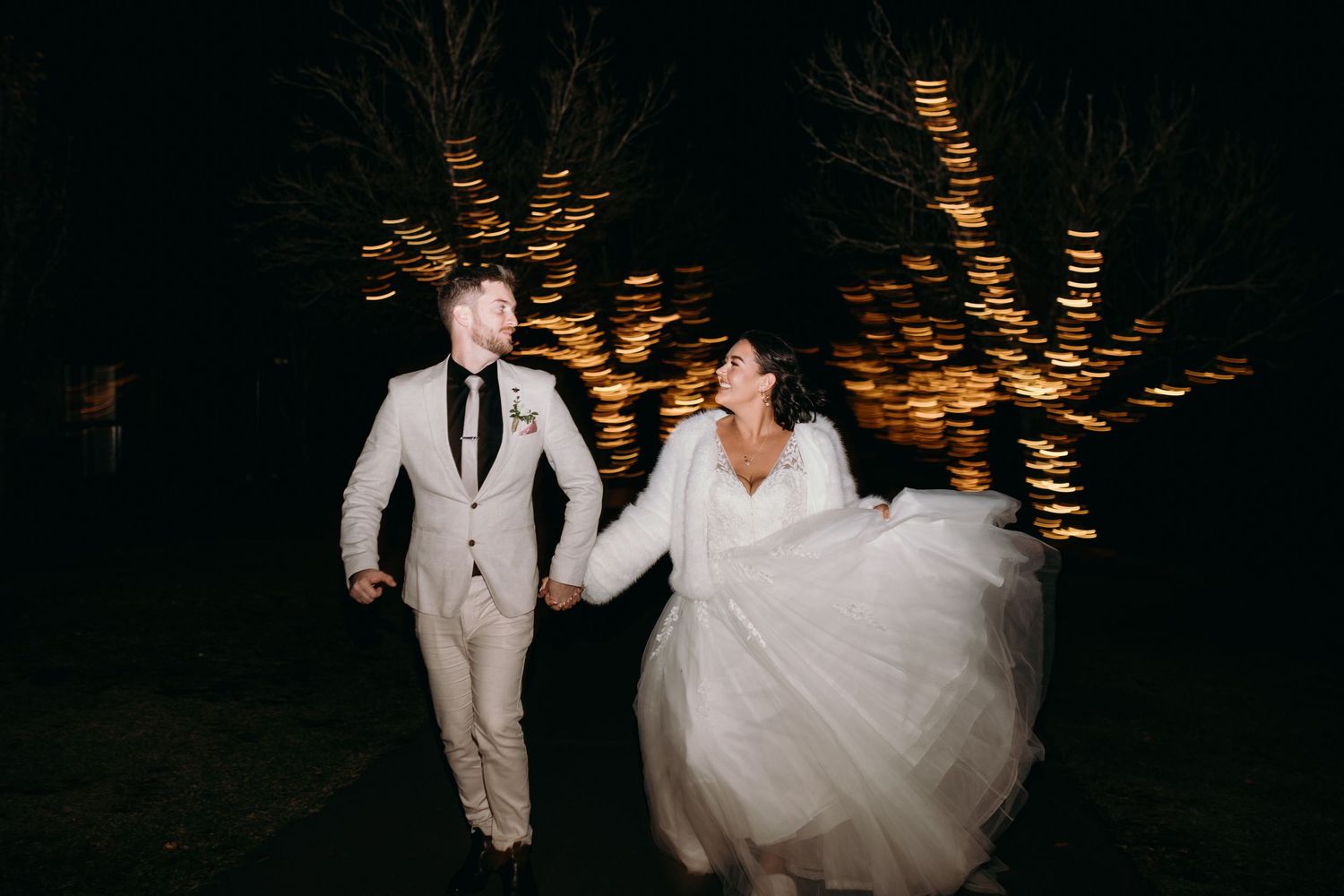 Couple runs through illuminated nighttime wedding venue with string lights overhead.