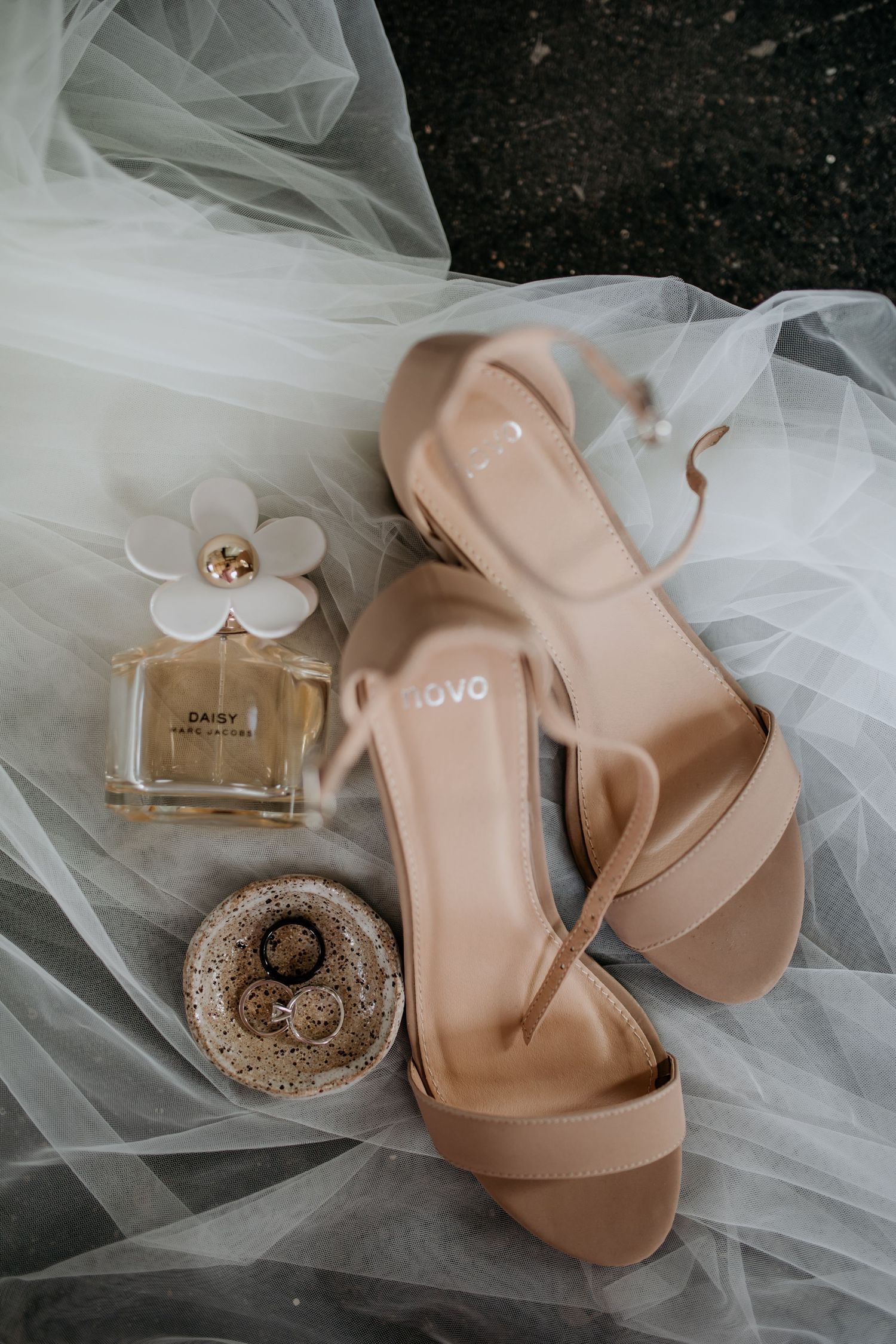 Wedding accessories including nude heels, perfume bottle and rings on delicate white tulle fabric.