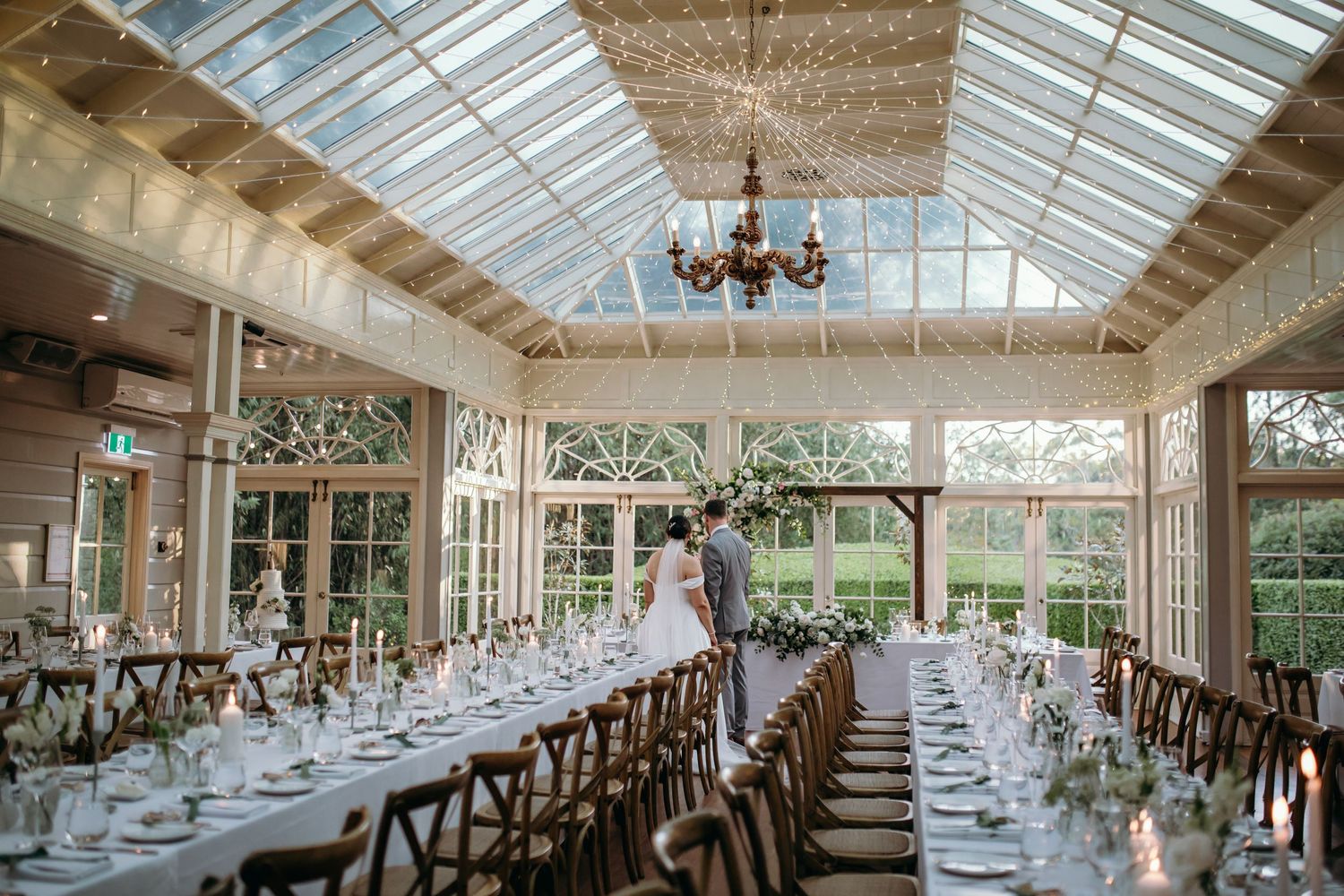 Elegant greenhouse wedding reception setup with long tables, white linens, and natural light streaming through glass roof.