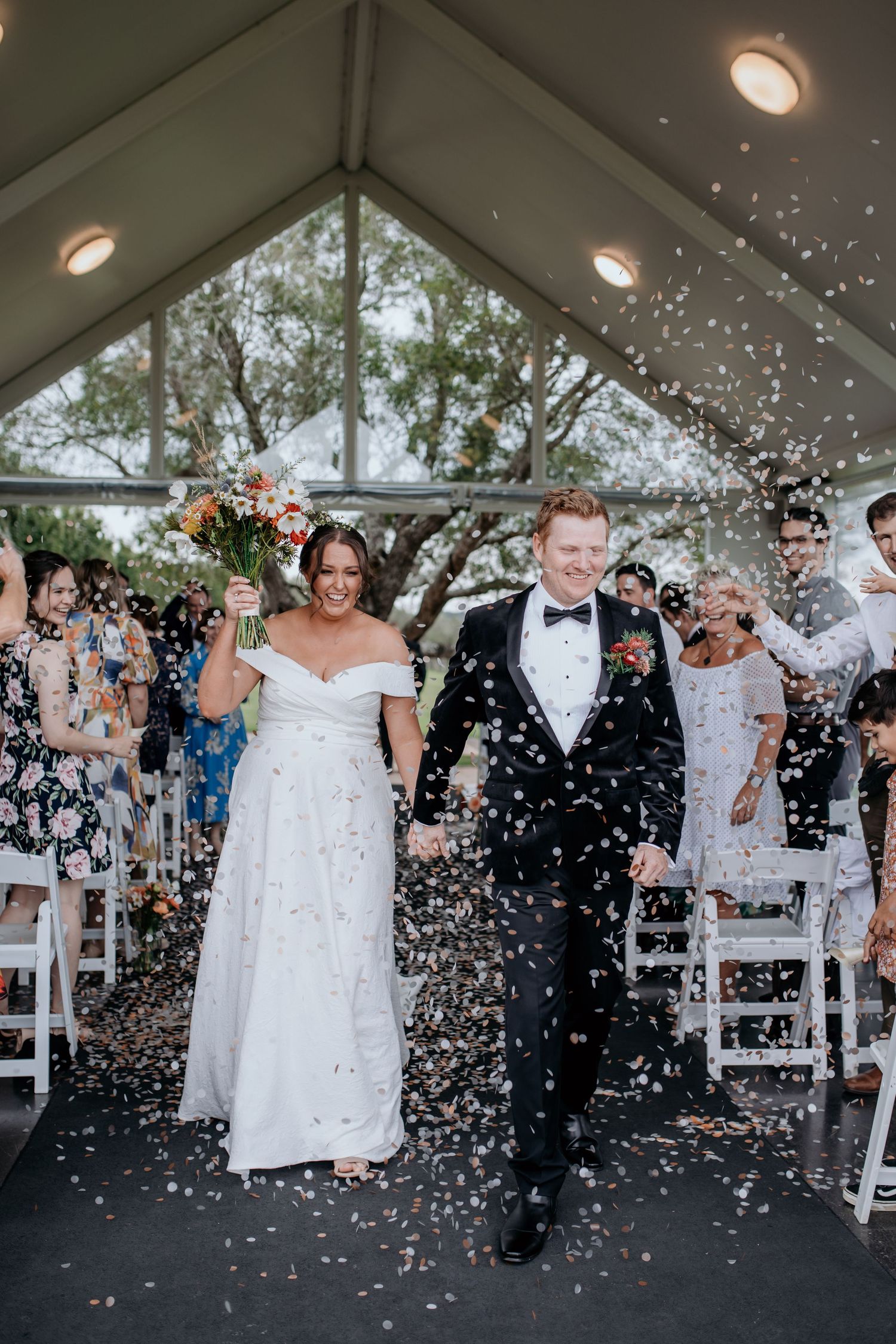 Wedding guests throw white confetti at a couple celebrating under a clear tent during an outdoor ceremony.