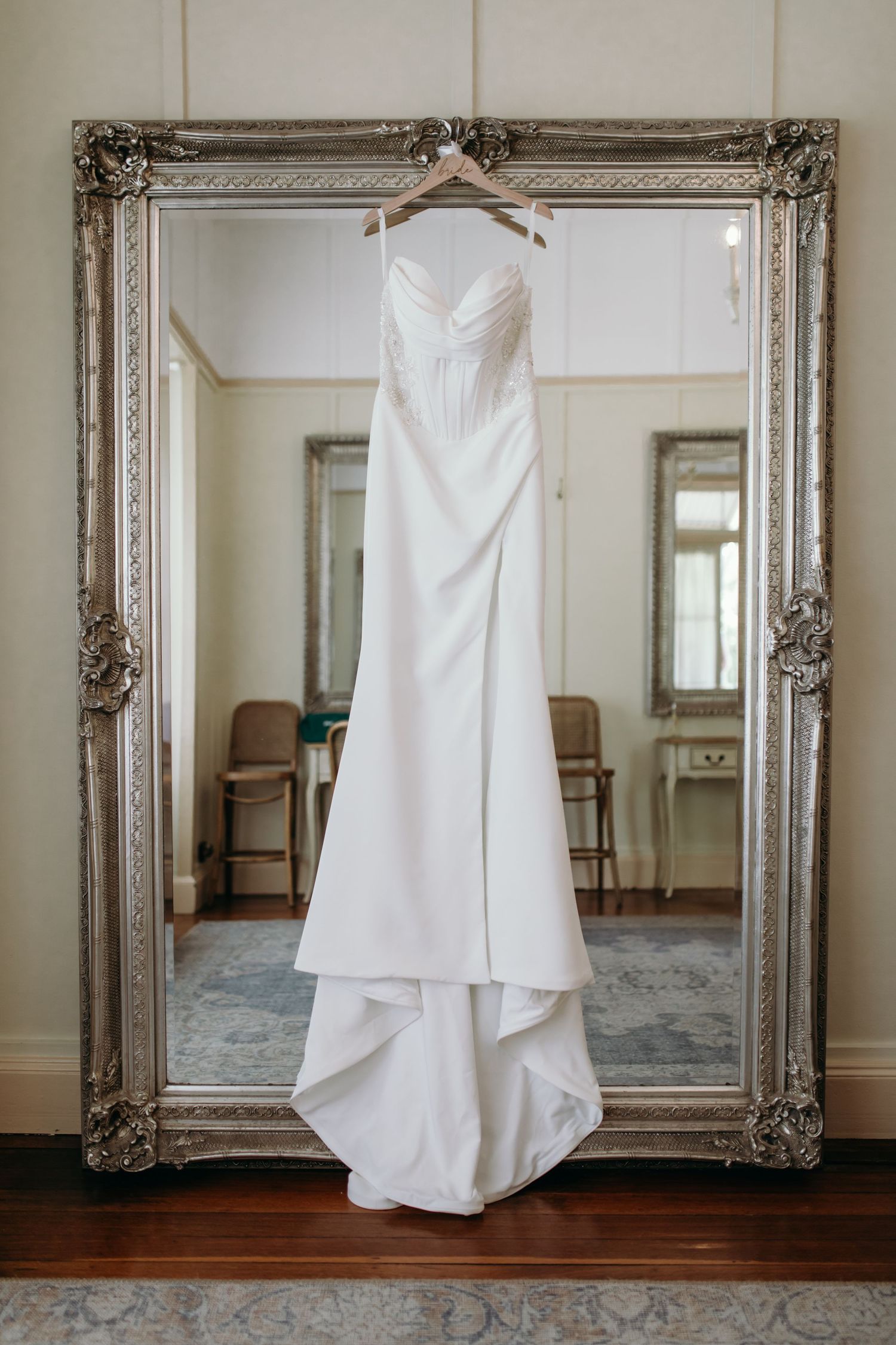 A white wedding dress hangs elegantly in front of an ornate silver-framed mirror against a neutral wall.