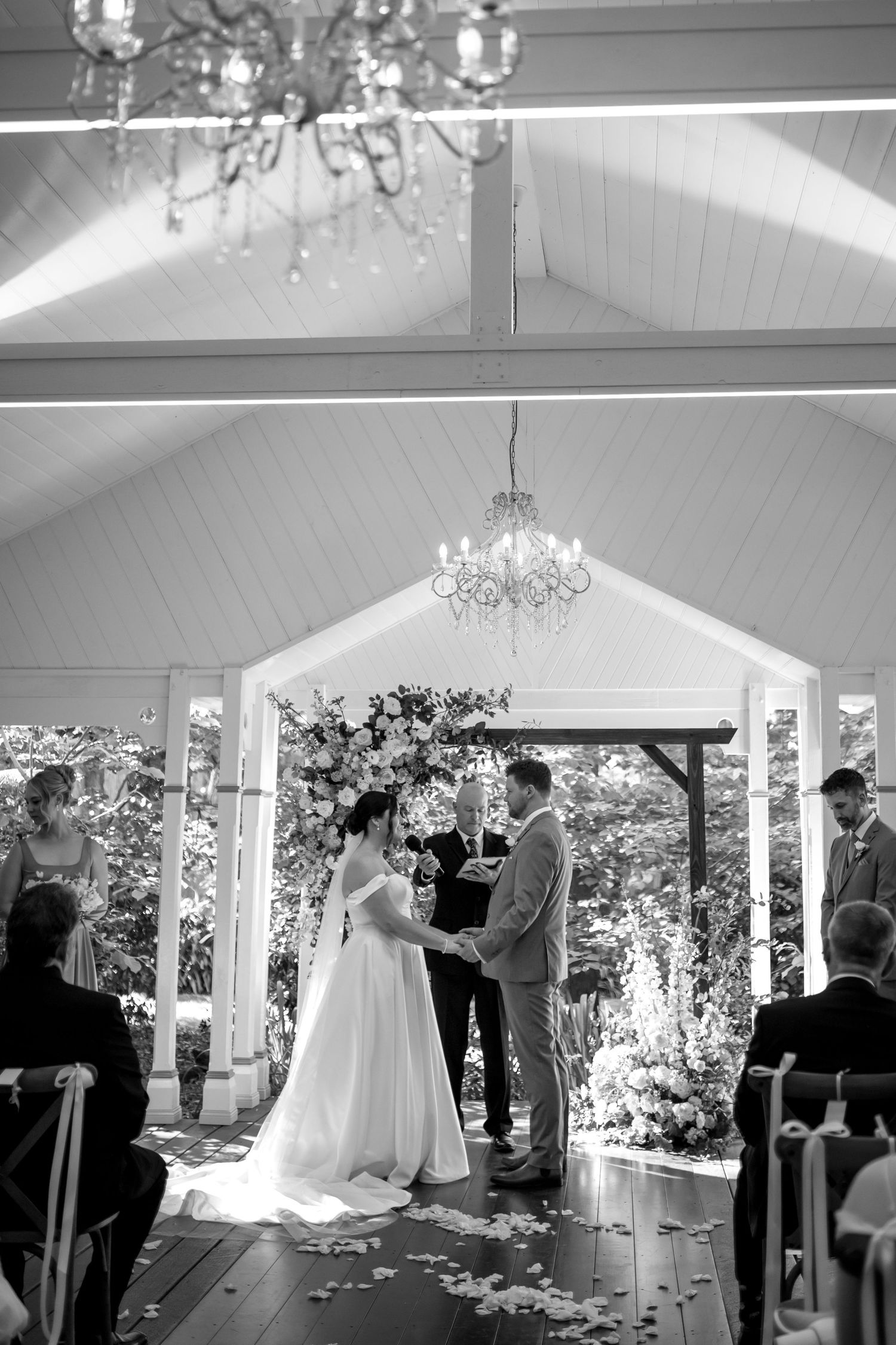 A wedding ceremony takes place in an elegant white pavilion with crystal chandeliers and natural light streaming in.