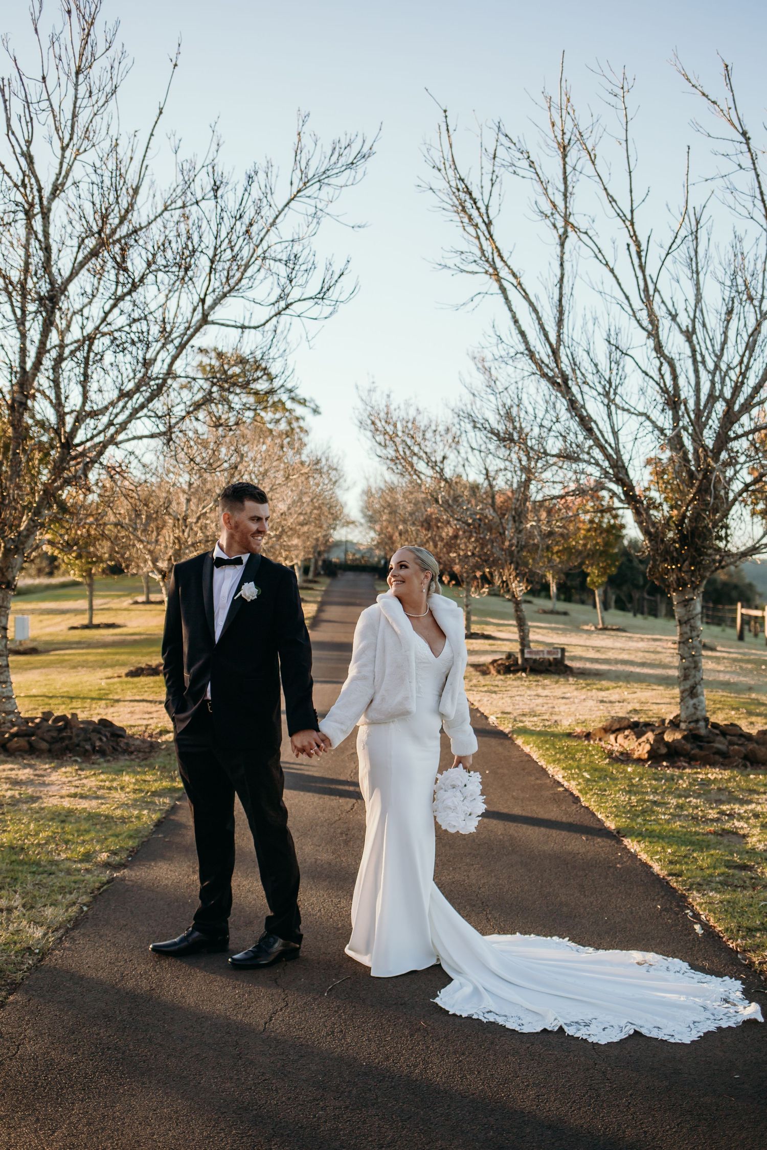 A couple in wedding attire walks along a winding path through a winter landscape with bare trees.