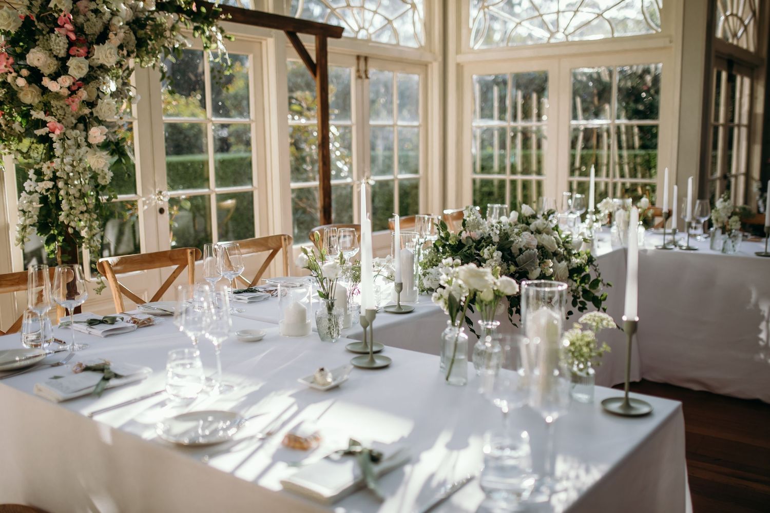 Elegant wedding reception tables set with white linens and floral centerpieces in a bright sunlit room.