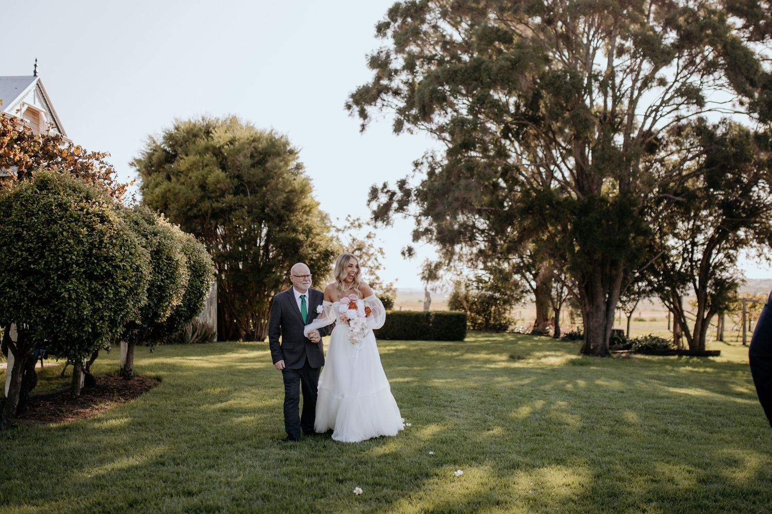 A scenic outdoor wedding ceremony set against a backdrop of trees and natural landscape. Bride is escorted by her father.