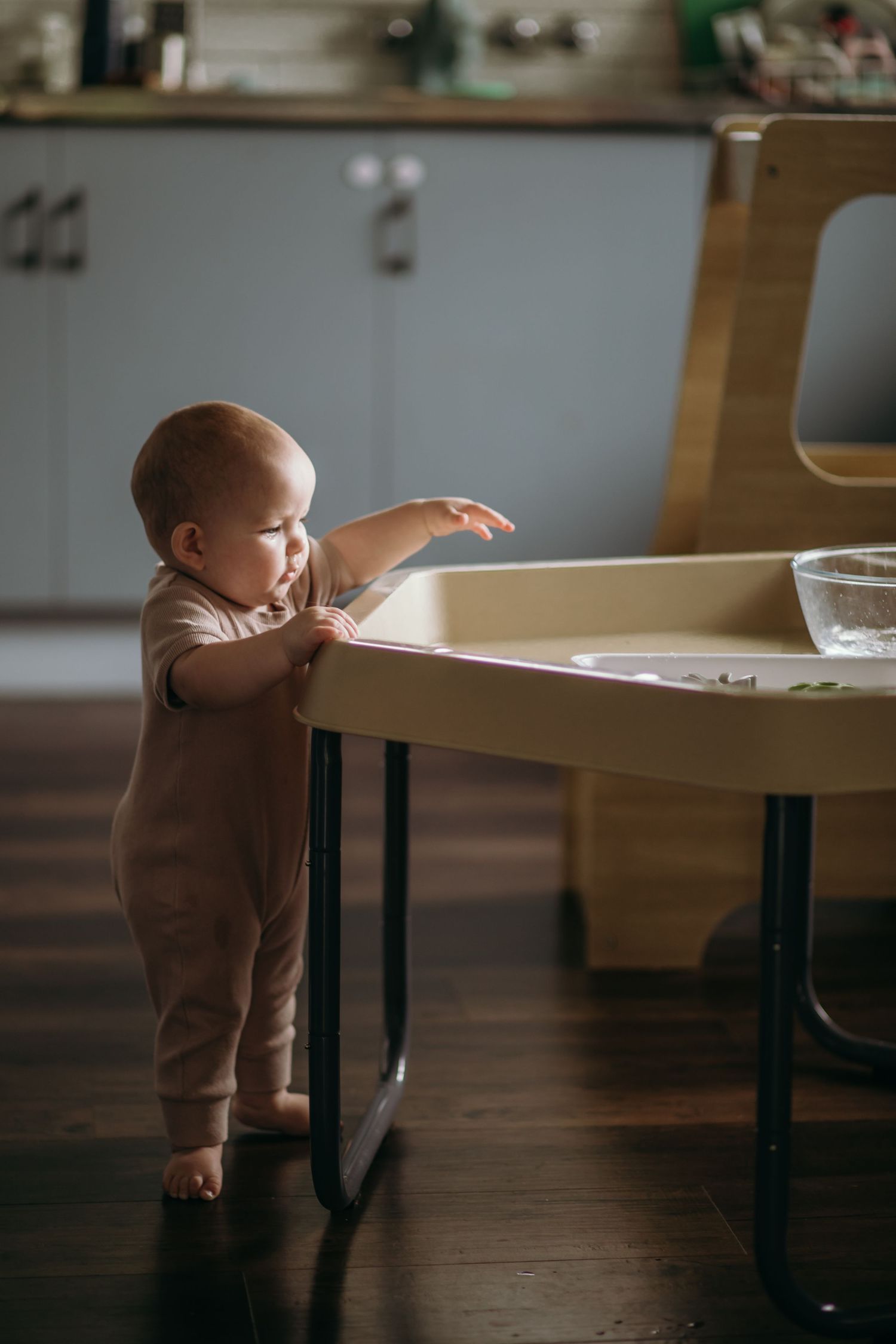 A toddler in brown clothing exploring a wooden table in a minimalist room.